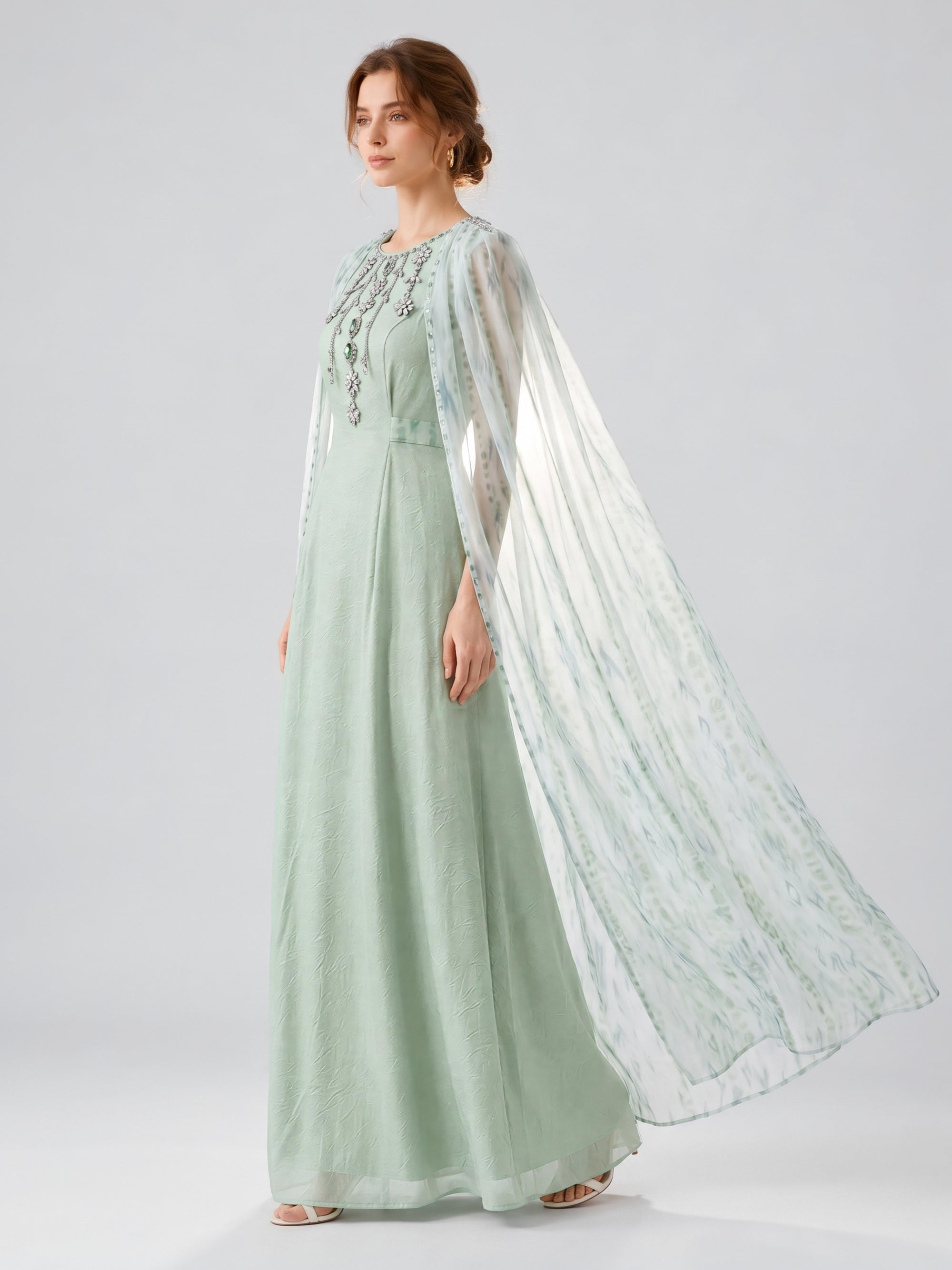 A woman, shown in profile against a plain, light background, wears the Social Queen Beaded Printed Cape Chiffon Dress—a floor-length, lightweight chiffon gown in light green with a sheer, flowing cape.