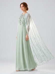 A woman, shown in profile against a plain, light background, wears the Social Queen Beaded Printed Cape Chiffon Dress—a floor-length, lightweight chiffon gown in light green with a sheer, flowing cape.