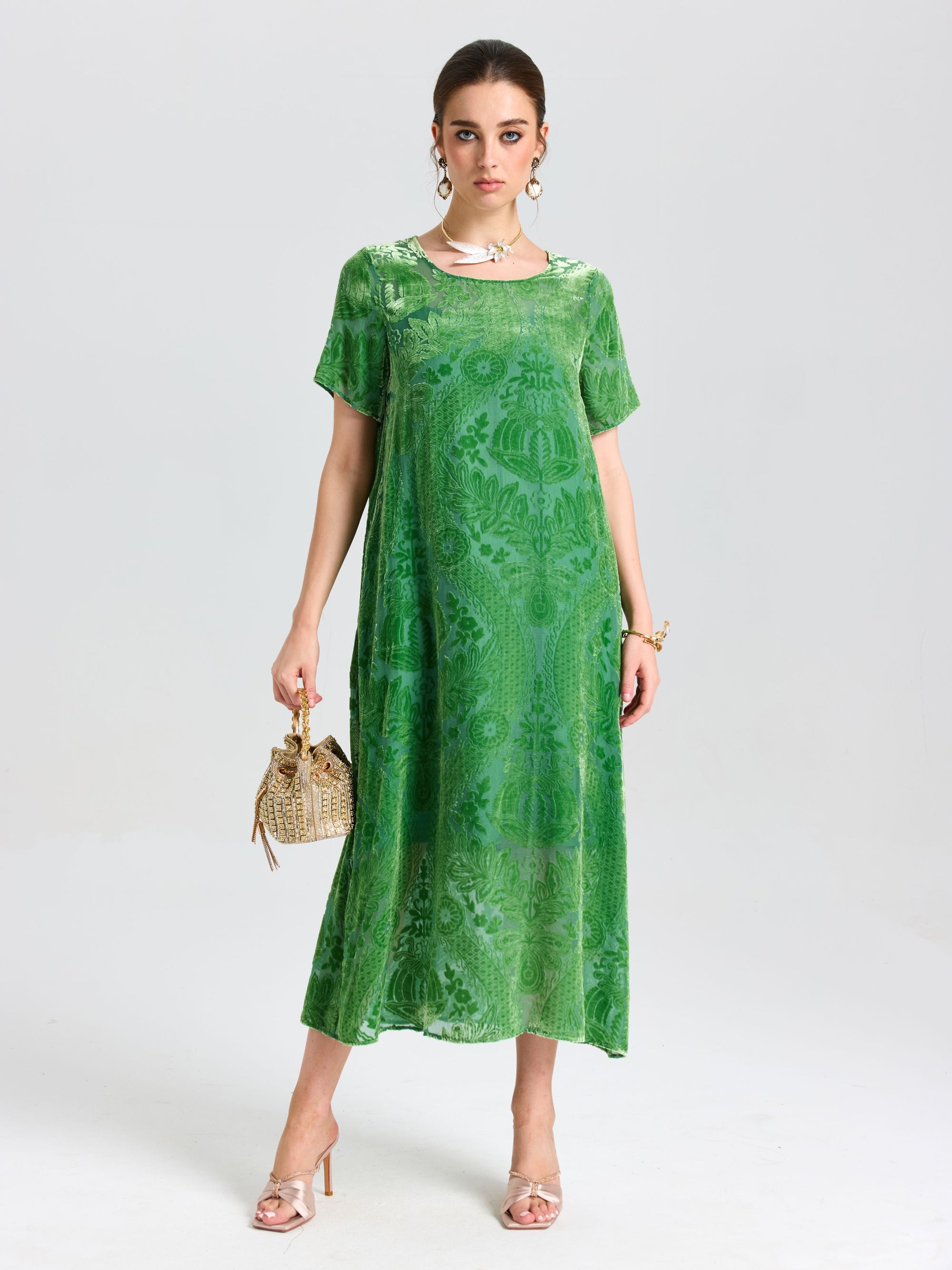 A woman stands against a plain background in the Social Queen Floral Silk Velvet Shift Dress (Cami Included), paired with metallic heels, a woven handbag, and styled with a statement necklace and bracelet.