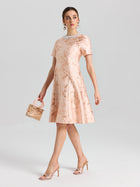 A woman wears the Social Queen Pearl Embellished Jacquard Dress, featuring a light pink floral pattern and pearl-trimmed neckline, paired with a small purse, heels, and statement earrings.