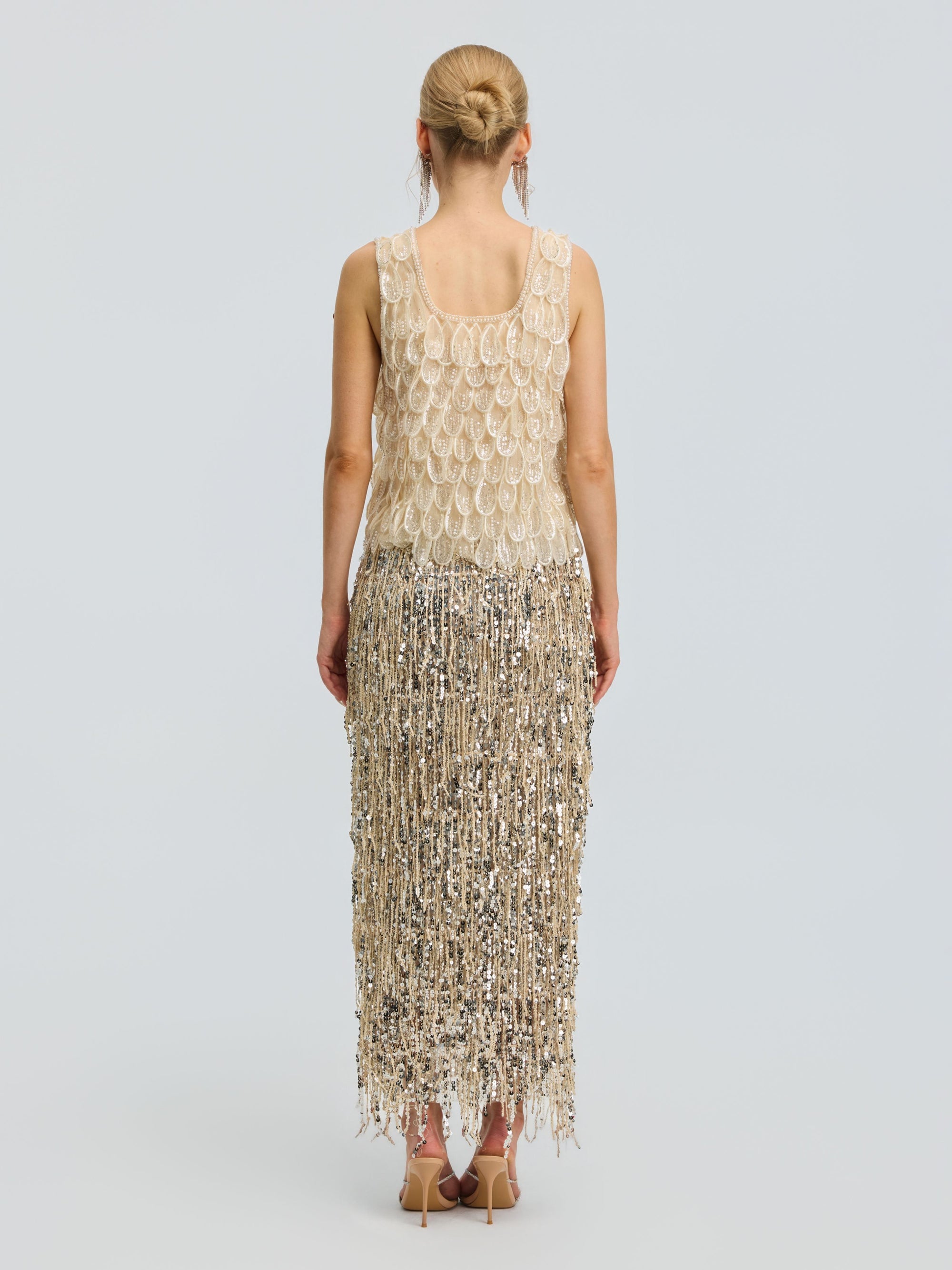 A woman stands with her back to the camera, wearing the Social Queen 3D Leaf Beaded Mesh Top featuring gold and silver embellishments, fringe details, and delicate 3D leaf appliqués. She pairs it with nude heels, her hair styled in a neat bun, against a plain light gray background.
