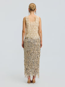A woman stands with her back to the camera, wearing the Social Queen 3D Leaf Beaded Mesh Top featuring gold and silver embellishments, fringe details, and delicate 3D leaf appliqués. She pairs it with nude heels, her hair styled in a neat bun, against a plain light gray background.