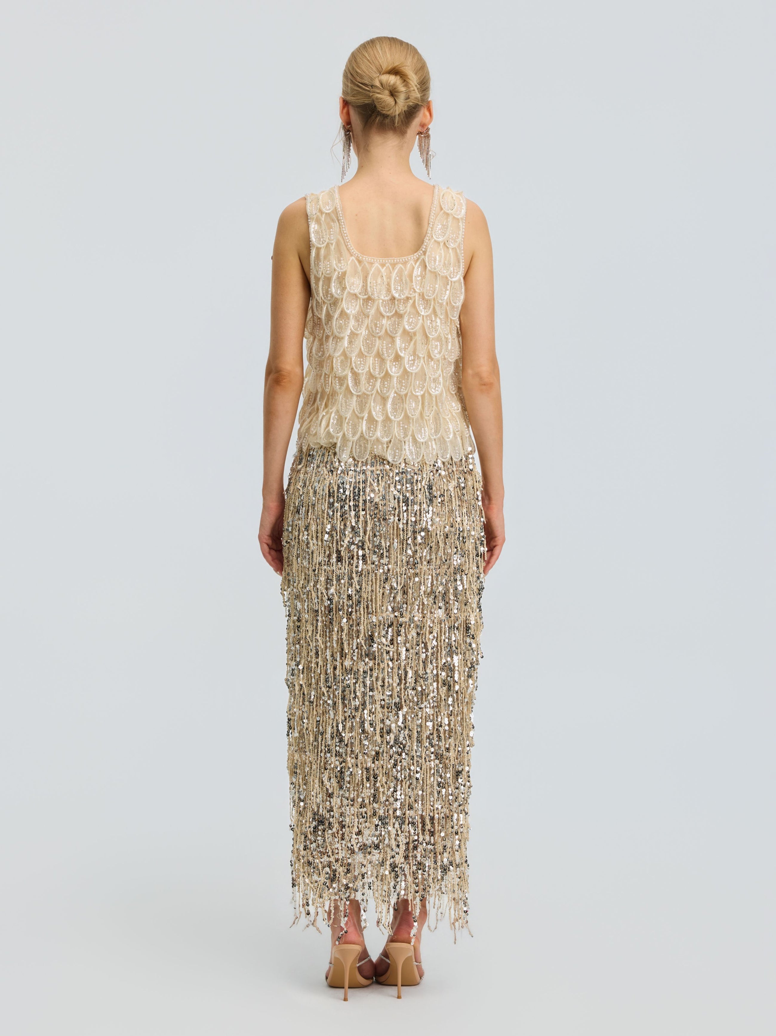 A woman stands with her back to the camera, wearing the Social Queen 3D Leaf Beaded Mesh Top featuring gold and silver embellishments, fringe details, and delicate 3D leaf appliqués. She pairs it with nude heels, her hair styled in a neat bun, against a plain light gray background.