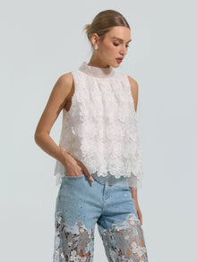 A woman is wearing the Social Queen 3D Floral Lace Appliqué Silk Top—a white sleeveless mulberry silk top adorned with delicate floral lace appliqué—paired with light blue jeans featuring lace cutout detailing, standing against a plain light background.