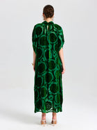 A person stands facing away in Social Queen's Floral Velvet Cape Dress (Cami Included), a long green silk velvet style with a cape-inspired silhouette, paired with beige heeled sandals against a plain white background.