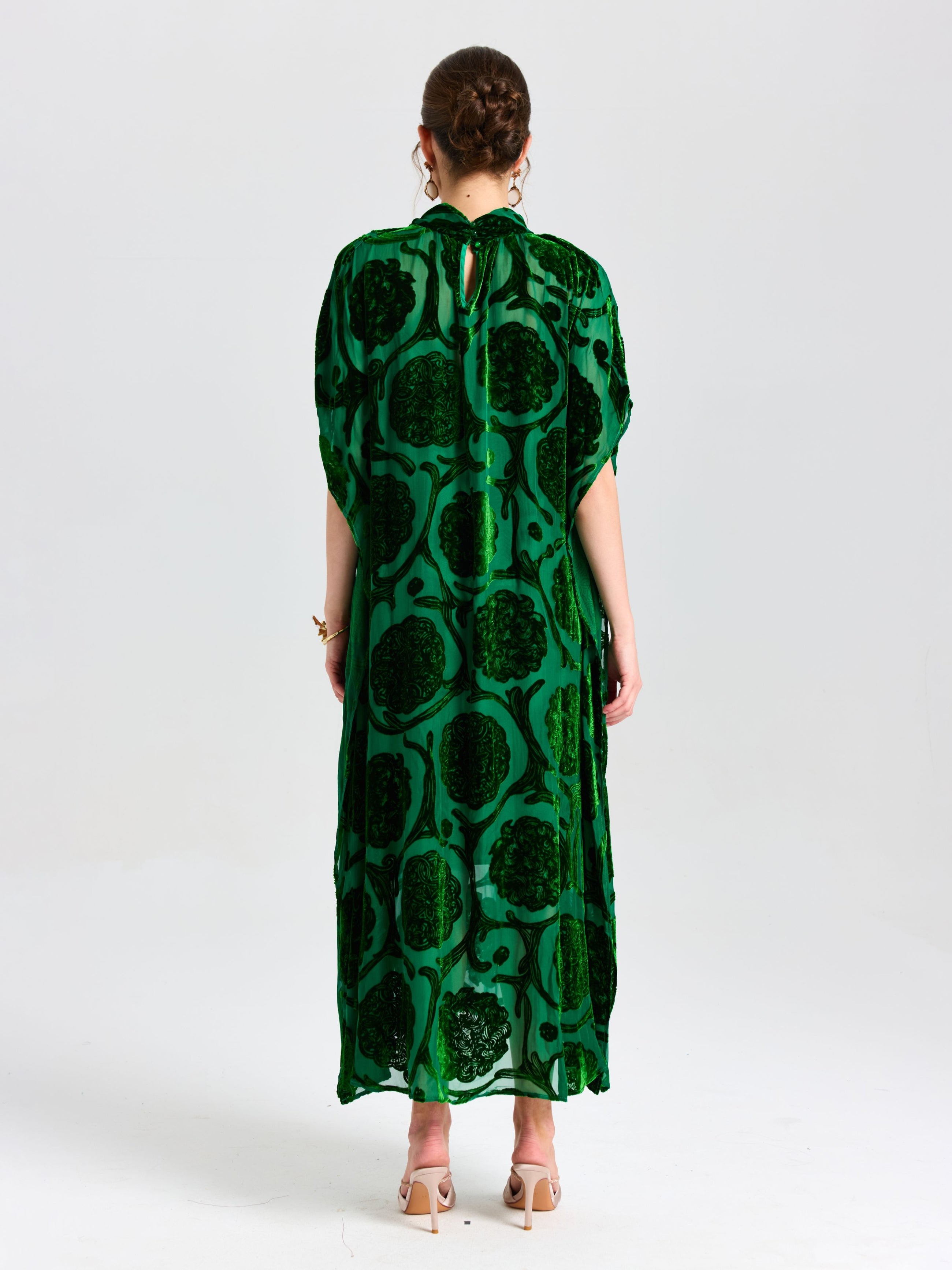 A person stands facing away in Social Queen's Floral Velvet Cape Dress (Cami Included), a long green silk velvet style with a cape-inspired silhouette, paired with beige heeled sandals against a plain white background.