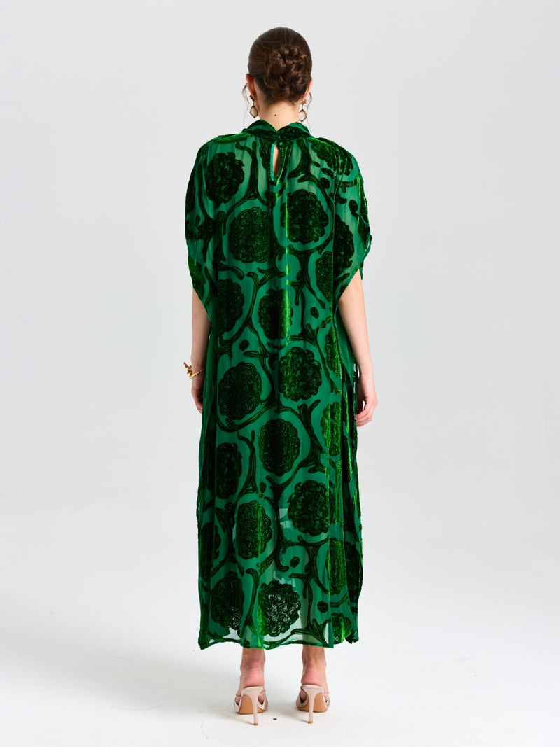 A person stands facing away in Social Queen's Floral Velvet Cape Dress (Cami Included), a long green silk velvet style with a cape-inspired silhouette, paired with beige heeled sandals against a plain white background.