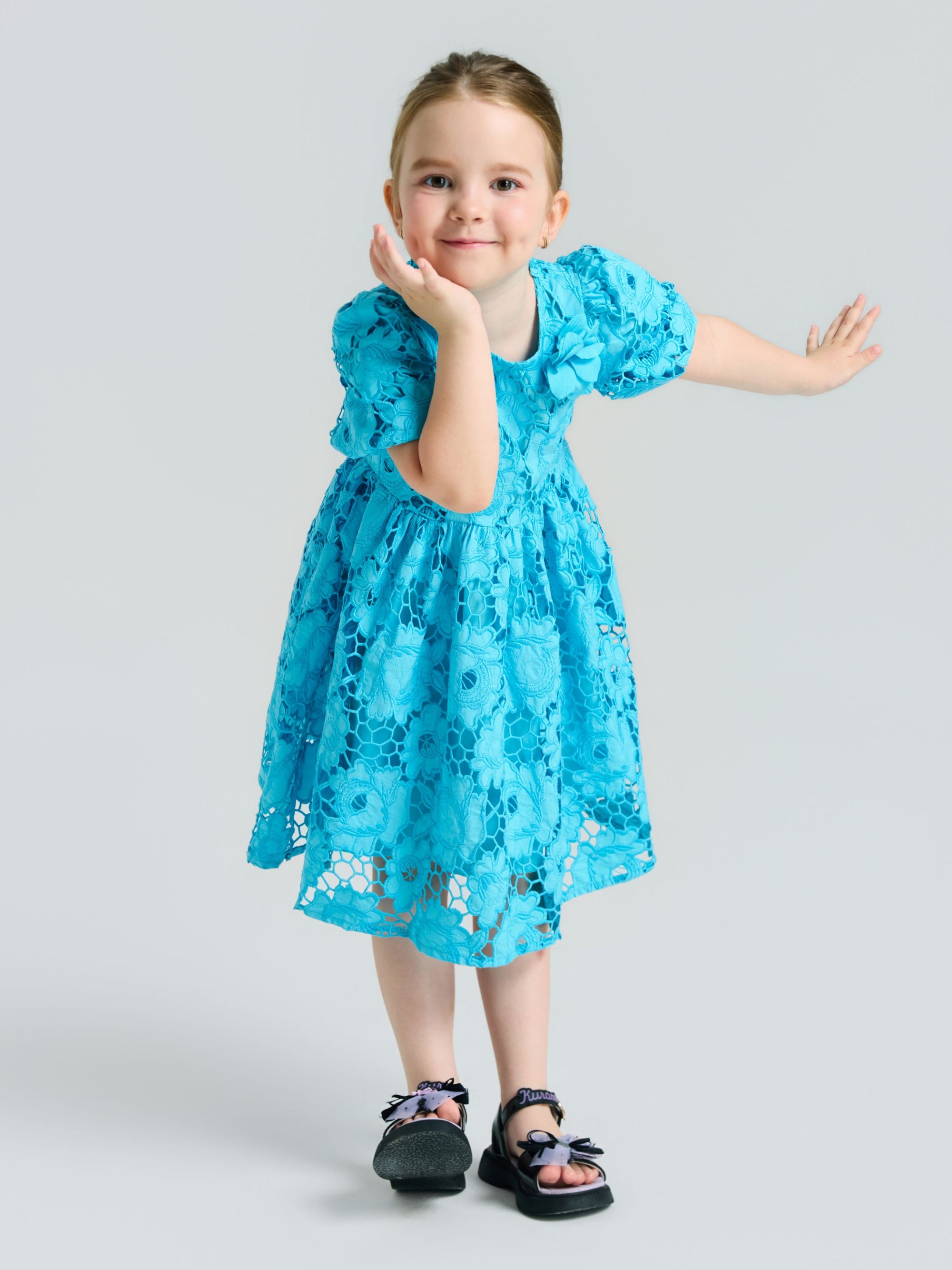 A young girl models the Social Queen Girls 3D Floral Appliqué Cutwork Lace Dress with black sandals, posing with one hand on her cheek and the other arm outstretched against a light gray background.