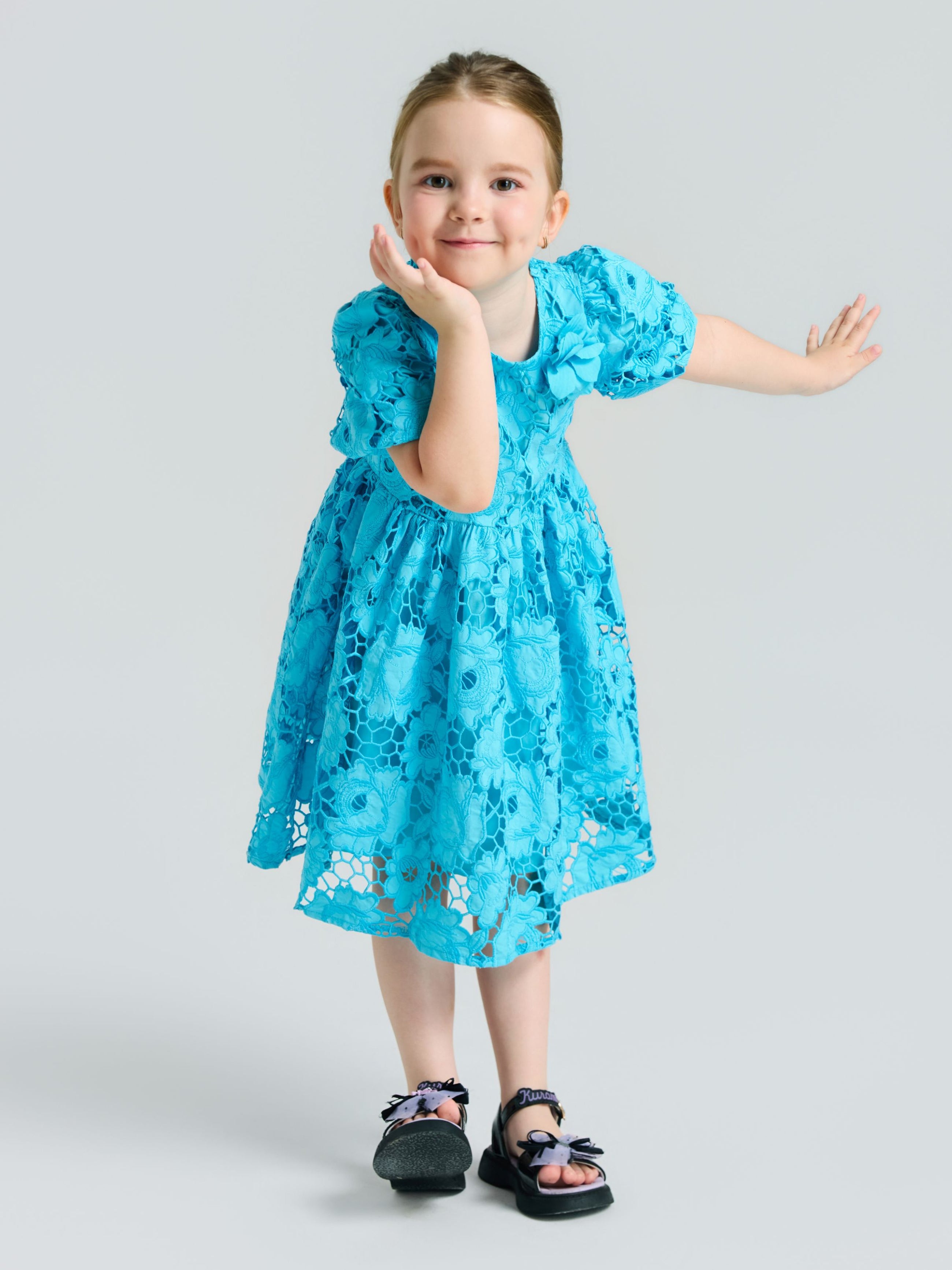 A young girl models the Social Queen Girls 3D Floral Appliqué Cutwork Lace Dress with black sandals, posing with one hand on her cheek and the other arm outstretched against a light gray background.