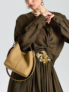 A woman wears a brown dress with a gold floral belt, carrying the Social Queen Woven Handle Pebbled Leather Bucket Bag over her shoulder. Her head is slightly turned and one hand rests near her chin.