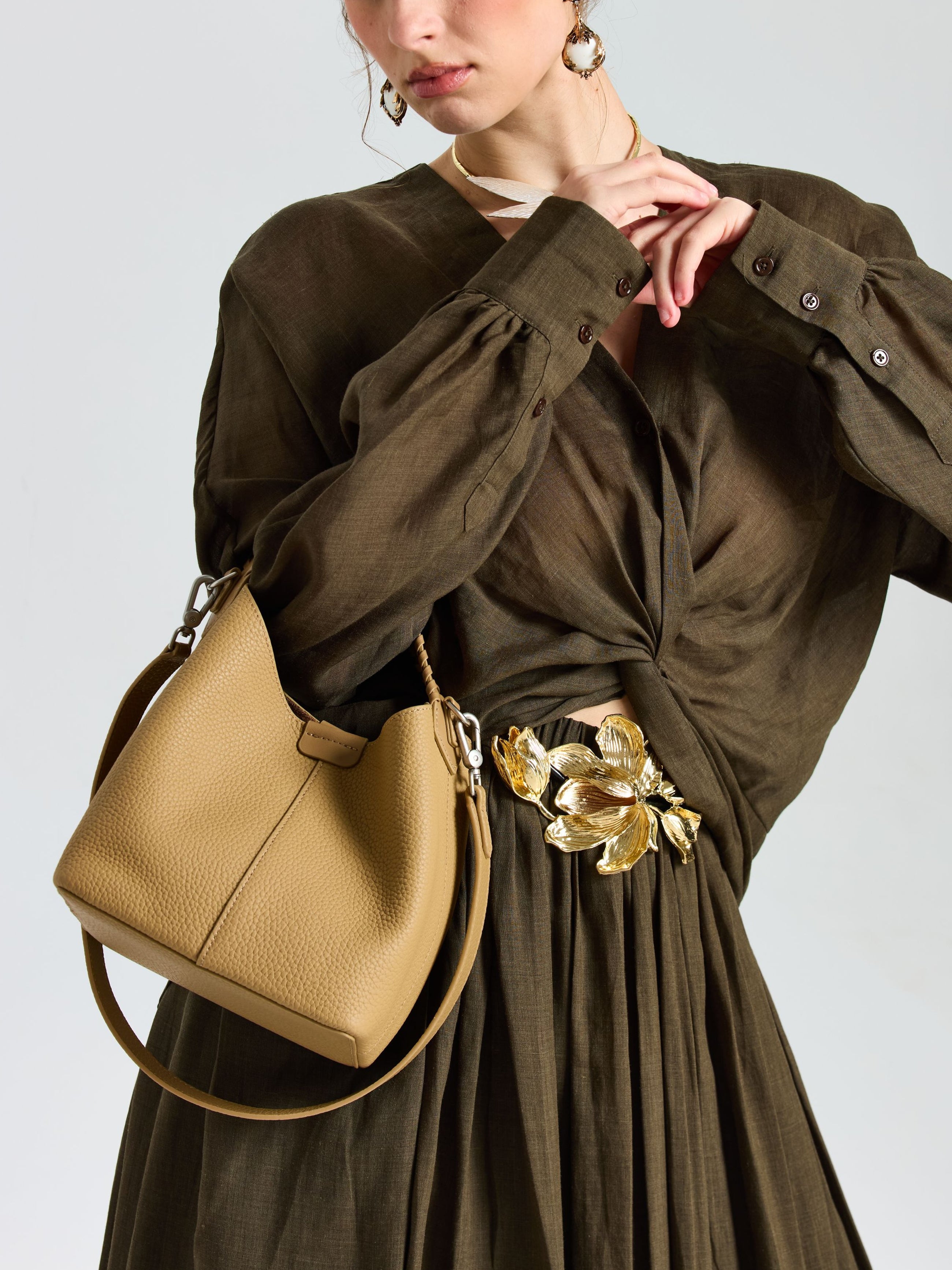 A woman wears a brown dress with a gold floral belt, carrying the Social Queen Woven Handle Pebbled Leather Bucket Bag over her shoulder. Her head is slightly turned and one hand rests near her chin.