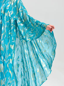 A model showcases the Gold Botanical Jacquard Silk Dress by Social Queen, highlighting its sheer turquoise and gold fabric and intricate botanical design against a simple background.