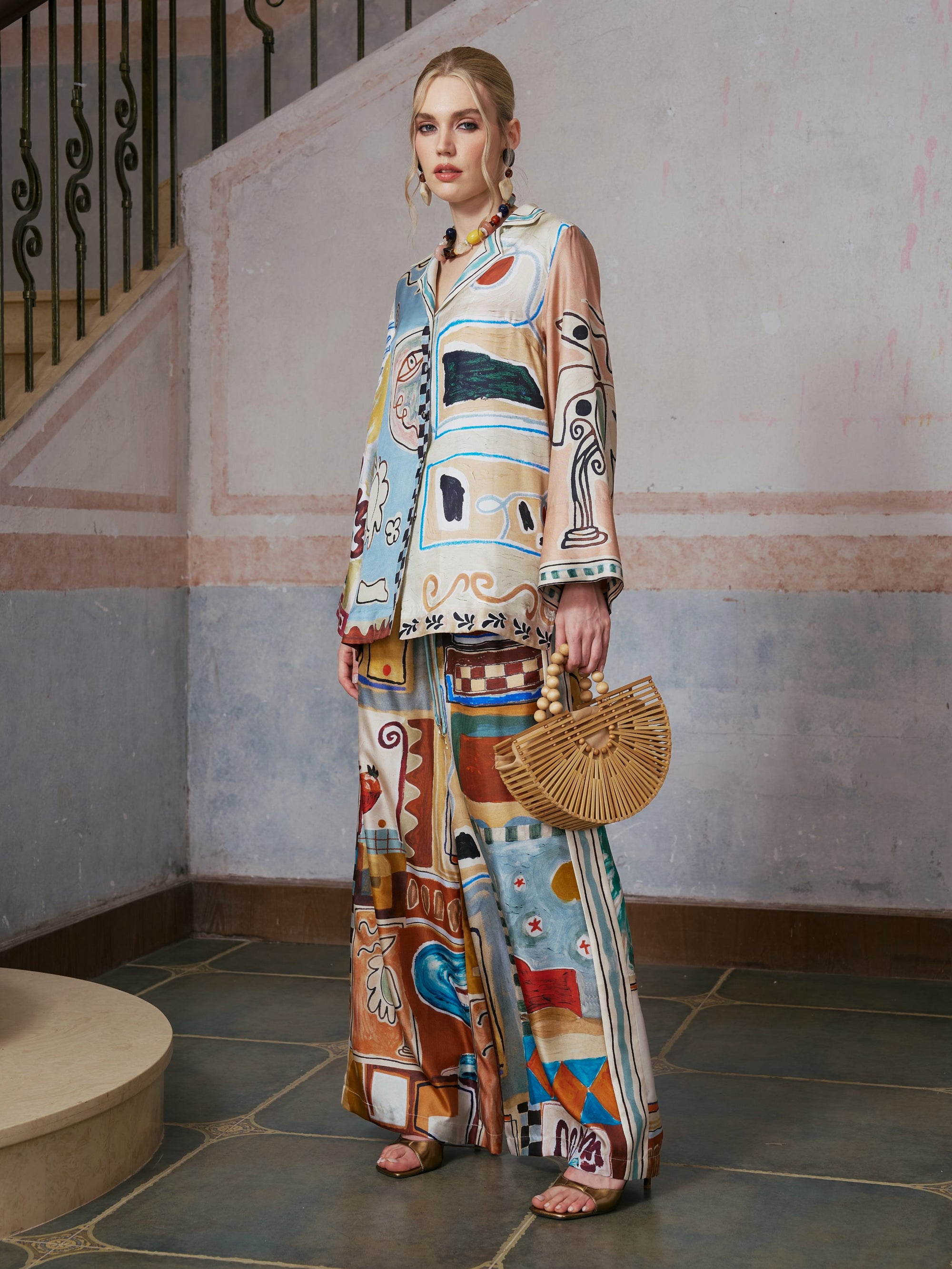 A woman stands indoors near a staircase with a wrought iron railing, holding a wooden fan-shaped bag and wearing wide-leg Abstract Print Mulberry Silk Pants by Social Queen, styled with a loose suit jacket featuring matching abstract patterns.