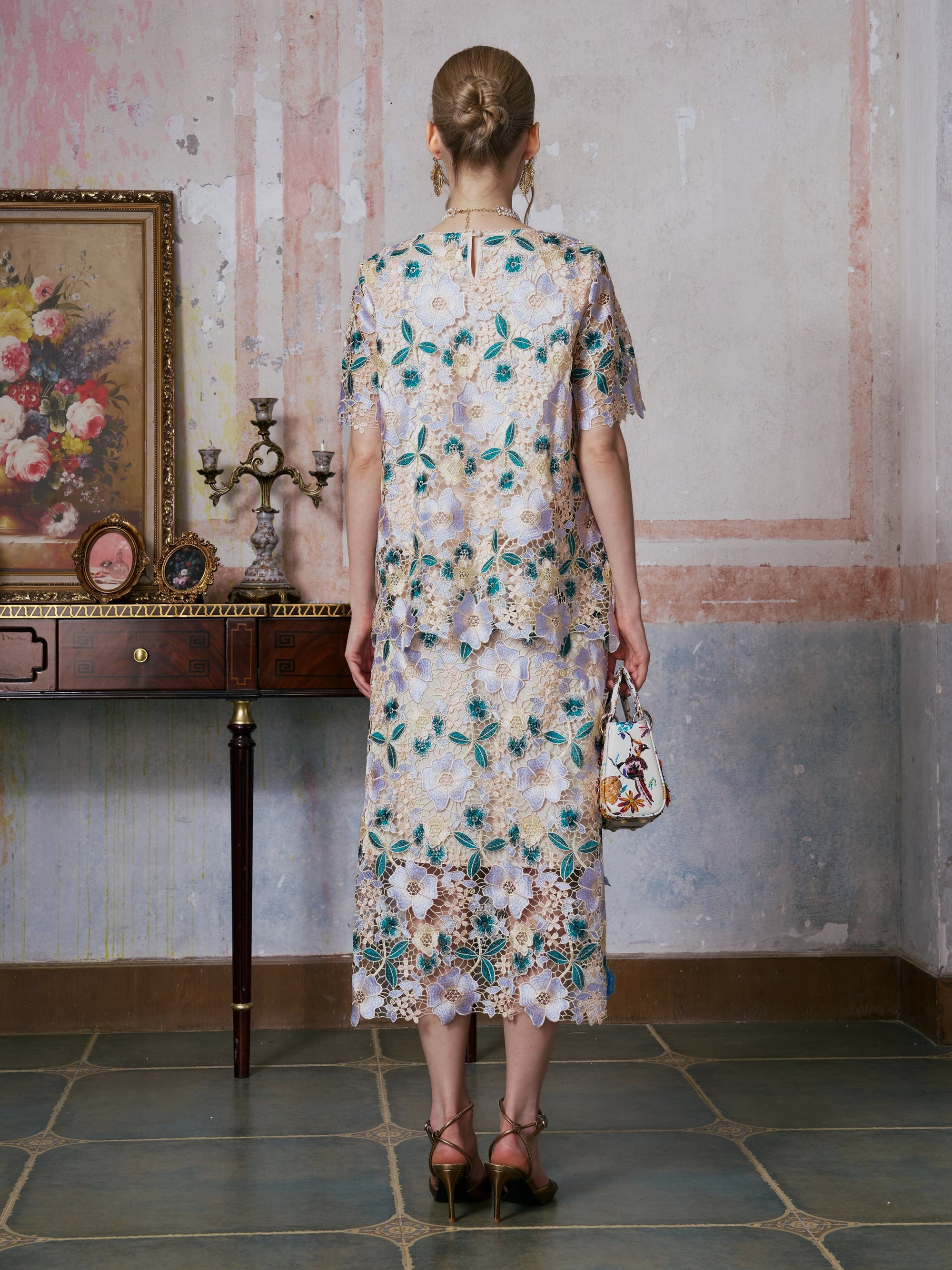 A person wearing the Social Queen 3D Floral Cutwork Lace Skirt and heels stands facing away, holding a small handbag in front of a vintage table adorned with a painting and candelabra.