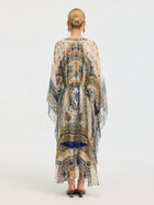 A woman faces away, wearing the Social Queen Byzantine Print Silk Caftan Dress with an intricate blue, gold, and white pattern. Her hair is styled in a neat bun and she completes the look with heeled sandals.