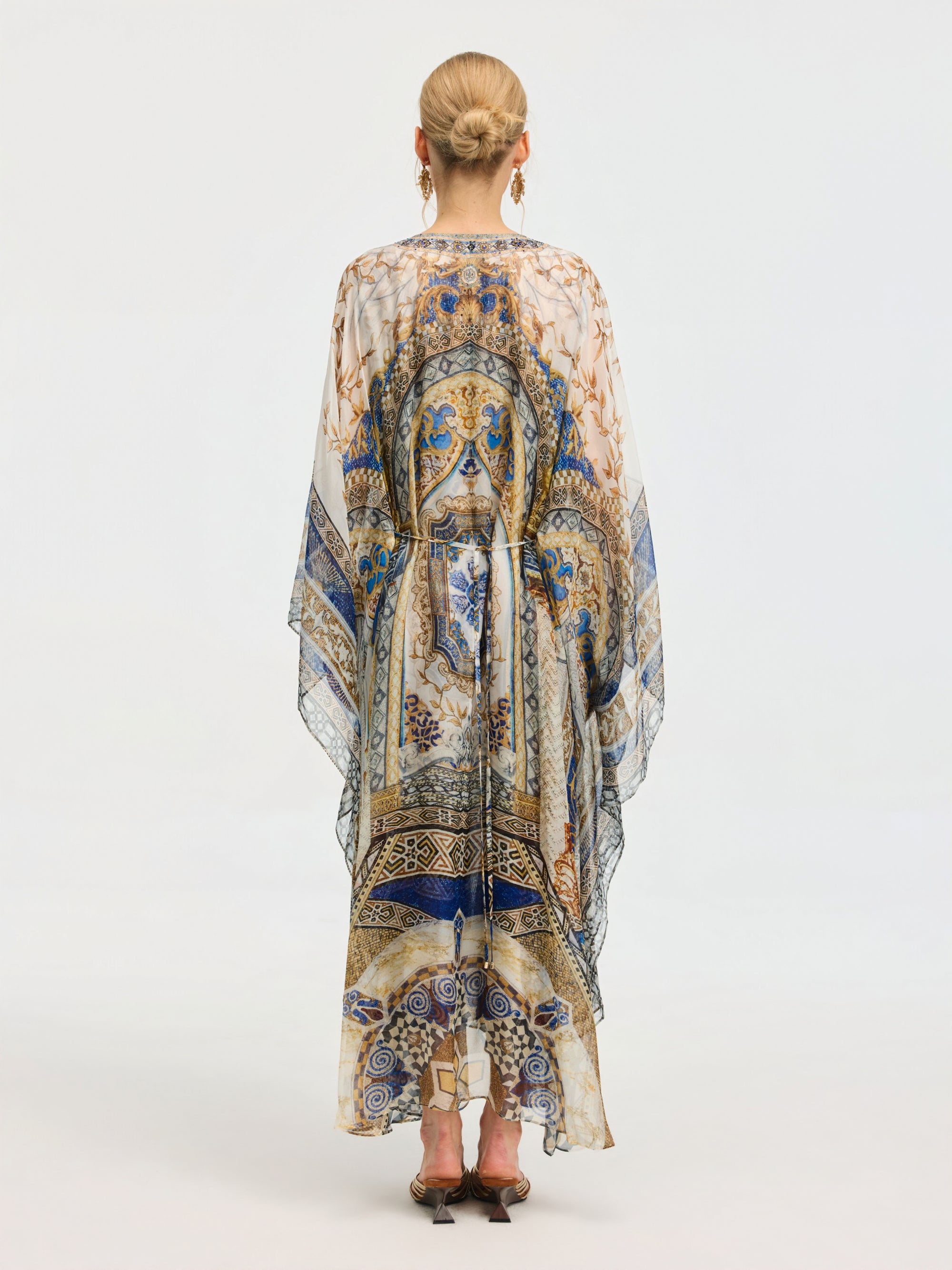 A woman faces away, wearing the Social Queen Byzantine Print Silk Caftan Dress with an intricate blue, gold, and white pattern. Her hair is styled in a neat bun and she completes the look with heeled sandals.