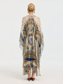 A woman faces away, wearing the Social Queen Byzantine Print Silk Caftan Dress with an intricate blue, gold, and white pattern. Her hair is styled in a neat bun and she completes the look with heeled sandals.