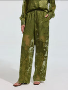Wearing Social Queen's Botanical Burnout Satin Wide-Leg Pants in olive green with a matching long-sleeve top and sandals, a person stands against a plain light background.