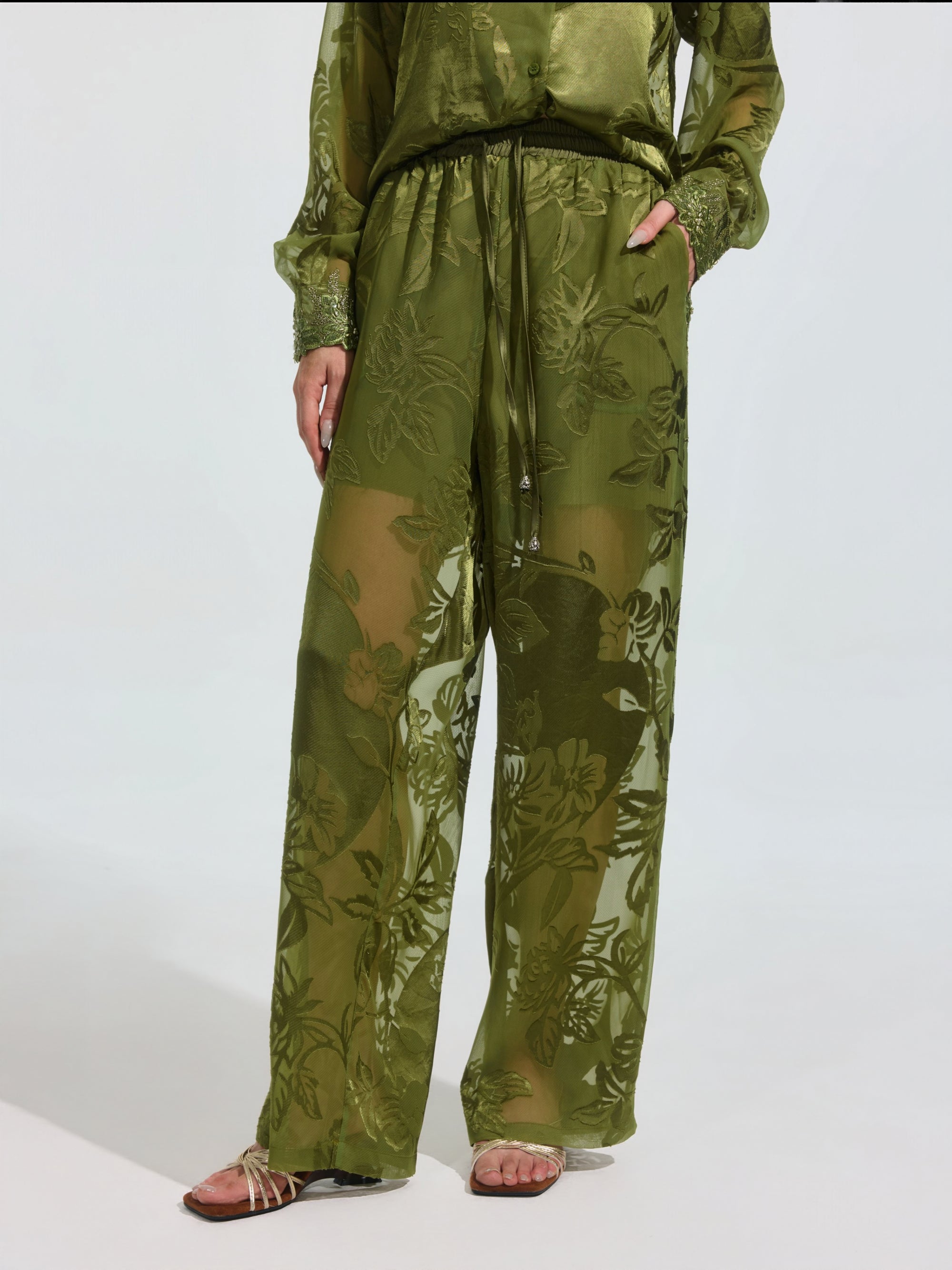 Wearing Social Queen's Botanical Burnout Satin Wide-Leg Pants in olive green with a matching long-sleeve top and sandals, a person stands against a plain light background.