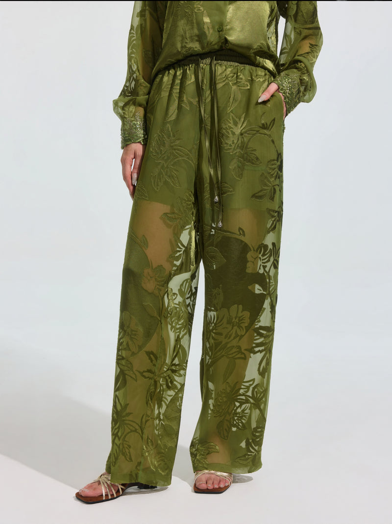 Wearing Social Queen's Botanical Burnout Satin Wide-Leg Pants in olive green with a matching long-sleeve top and sandals, a person stands against a plain light background.