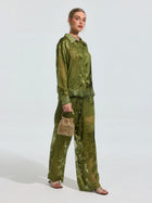 A woman wears the Social Queen Beaded Botanical Burnout Satin Shirt, featuring a green leaf pattern, styled with matching pants, a woven handbag, and flat sandals.