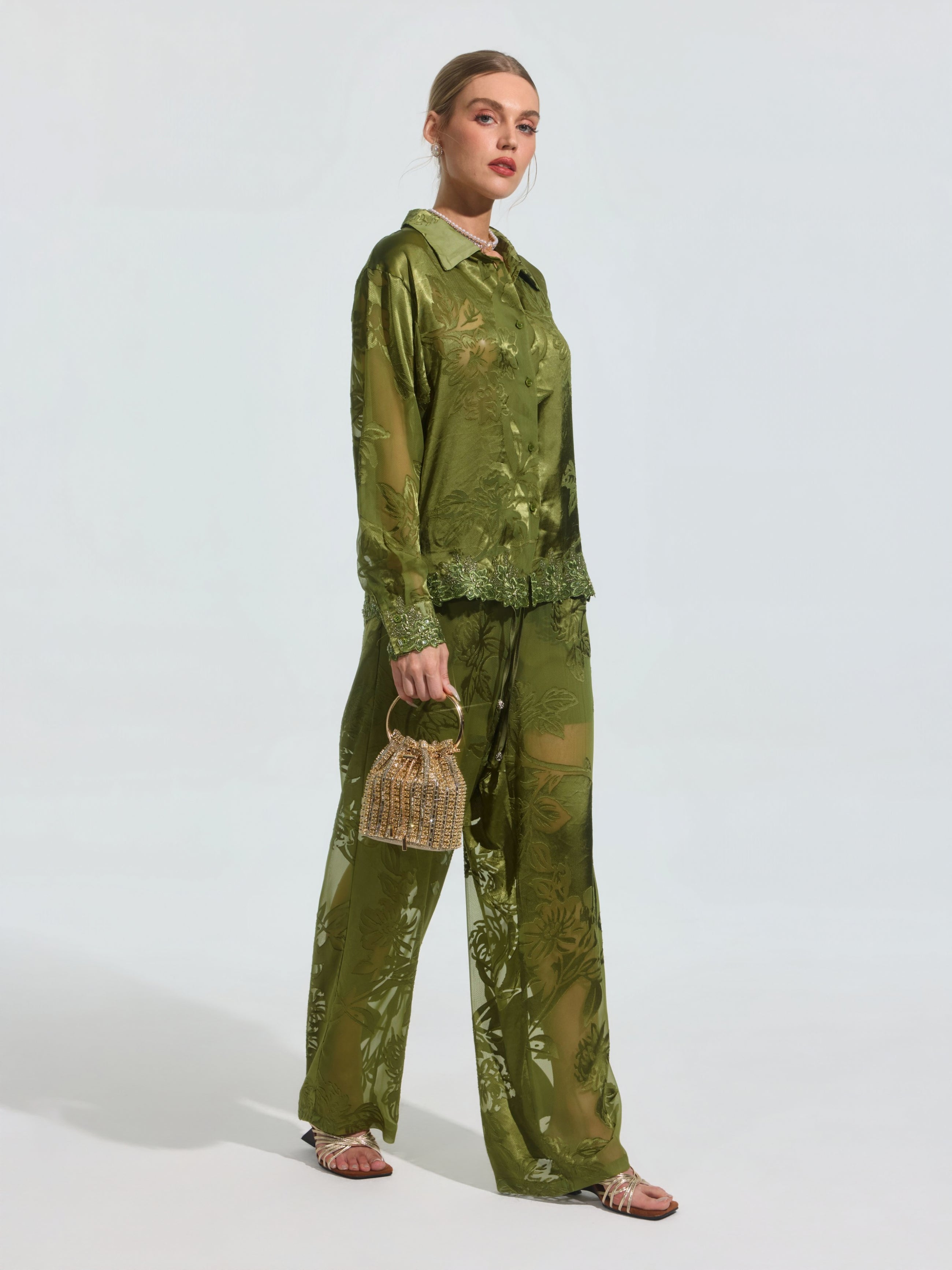 A woman wears the Social Queen Beaded Botanical Burnout Satin Shirt, featuring a green leaf pattern, styled with matching pants, a woven handbag, and flat sandals.