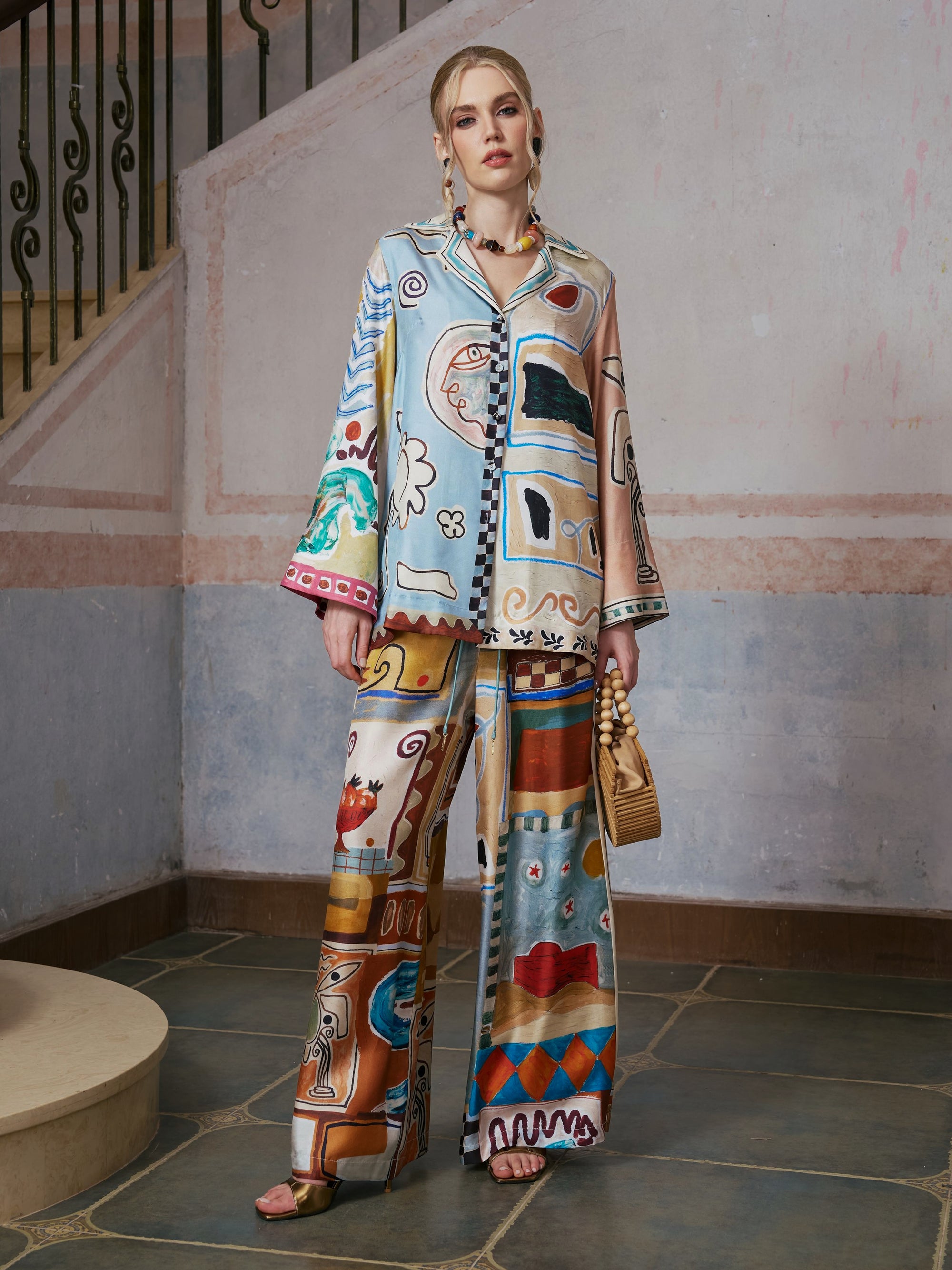 A woman stands indoors holding a small beige handbag, wearing Social Queen’s Abstract Print Mulberry Silk Pants—a matching set with colorful abstract patterns—set against a staircase and textured wall background.