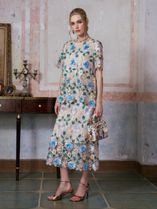 A woman stands indoors in front of a textured wall, wearing the Social Queen 3D Floral Cutwork Lace Skirt paired with a floral lace dress featuring blue flowers. She holds a small handbag and completes her look with gold sandals.
