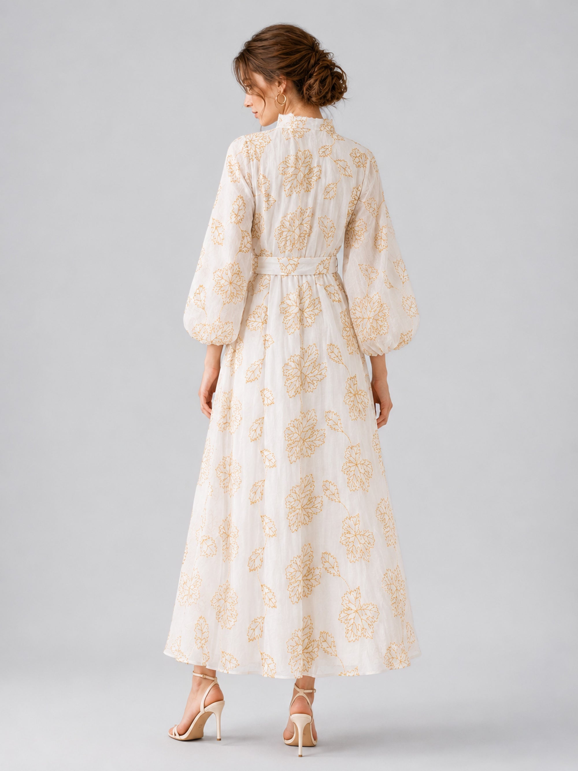 A woman stands with her back to the camera, modeling the Social Queen Leaf Embroidered Beaded Dress, a long, linen-like gown featuring gold leaf embroidery, long sleeves, a belted waist, and paired with high heels against a plain gray background.