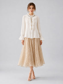 A woman is standing against a plain background wearing the Social Queen Pearl Embellished Ruffle Collar Blouse, a beige lace midi skirt, and nude heels.
