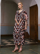 A woman stands indoors wearing the Social Queen Geometric Motif Print Silk Dress (Scarf Included), a patterned, loose-fitting mulberry silk dress with relaxed batwing sleeves. She holds a brown handbag and wears gold sandals, set against a backdrop of tiled floor and a wooden door.