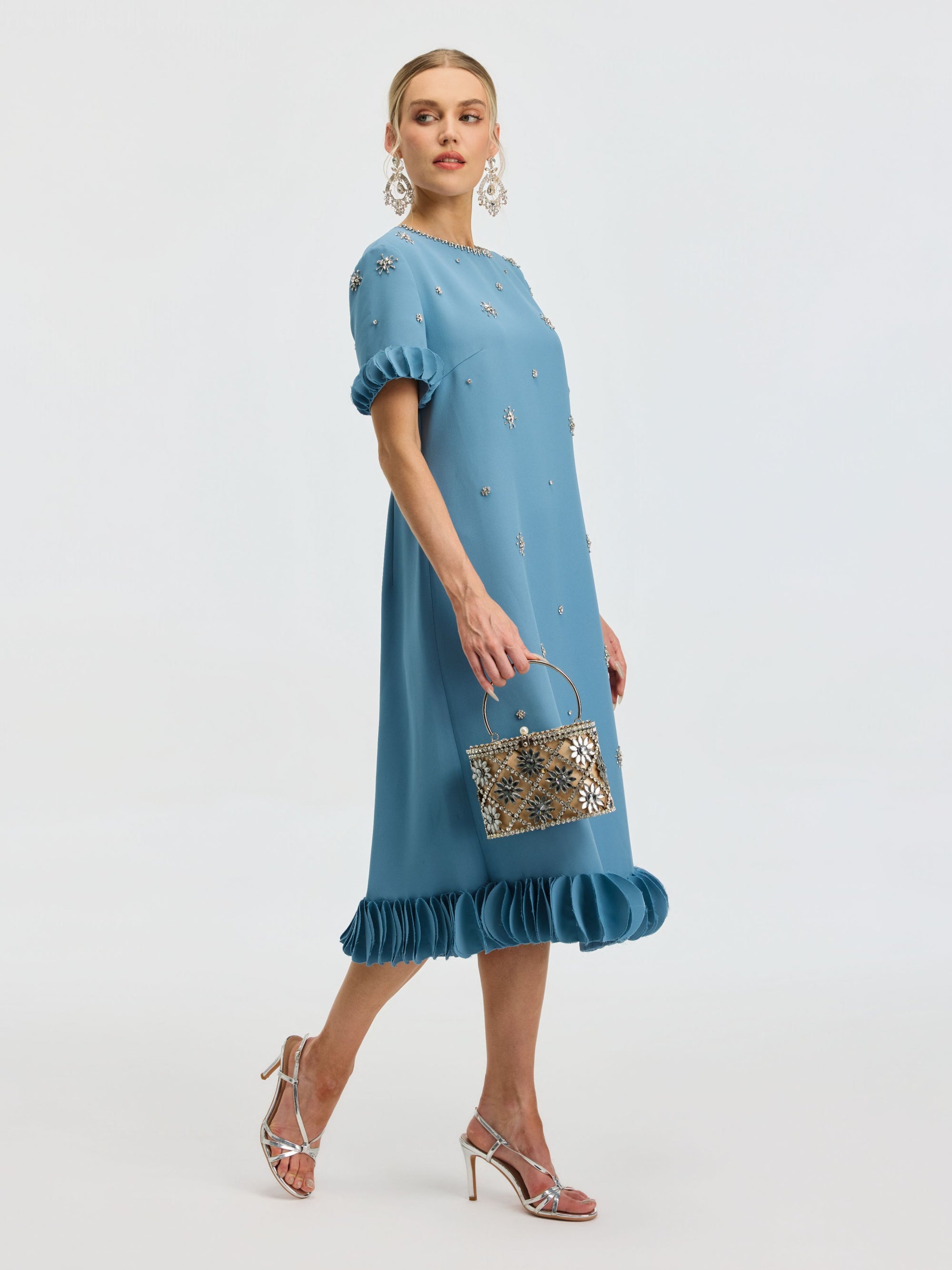 A woman models the Social Queen Beaded Crystal Embellished Dress, featuring a ruffle hem and beaded neckline, paired with a silver handbag, matching heels, and bold earrings.