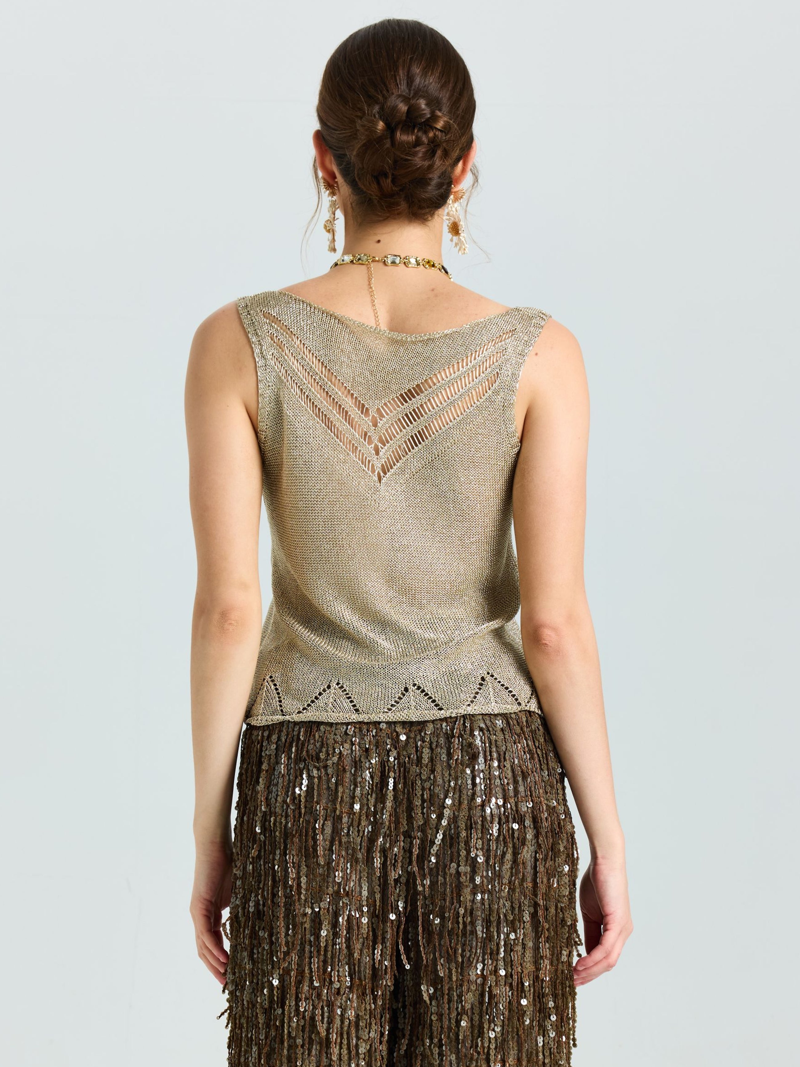 A woman with her hair in a neat bun faces away, wearing the Social Queen Open-Knit Metallic Sleeveless Top paired with a brown sequined skirt featuring geometric cutouts.