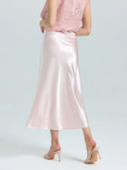 A person wears a light pink lace top, Social Queen's Satin A-Line Midi Skirt in shiny pink, and nude heeled sandals, standing against a plain light background.
