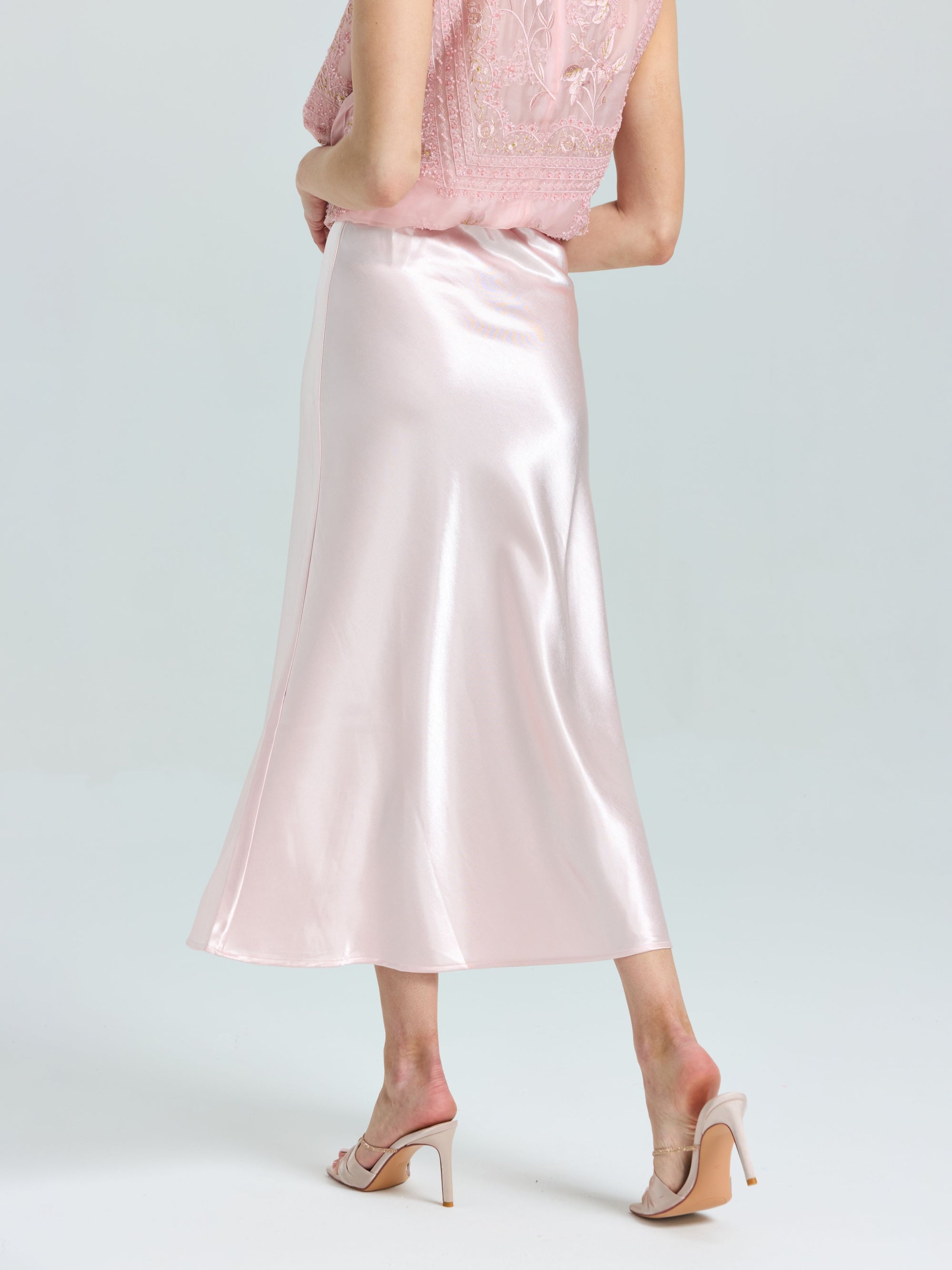 A person wears a light pink lace top, Social Queen's Satin A-Line Midi Skirt in shiny pink, and nude heeled sandals, standing against a plain light background.