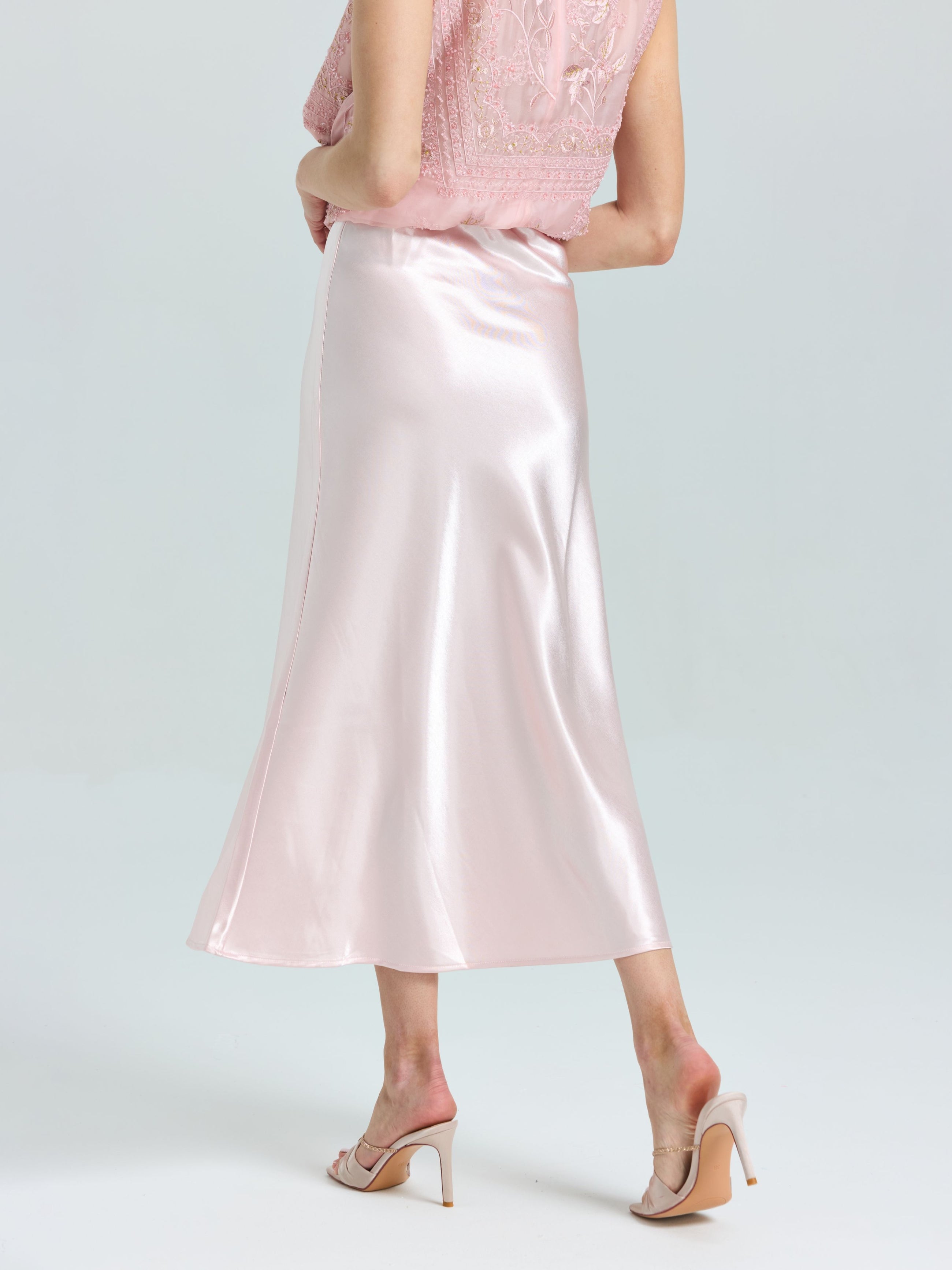 A person wears a light pink lace top, Social Queen's Satin A-Line Midi Skirt in shiny pink, and nude heeled sandals, standing against a plain light background.