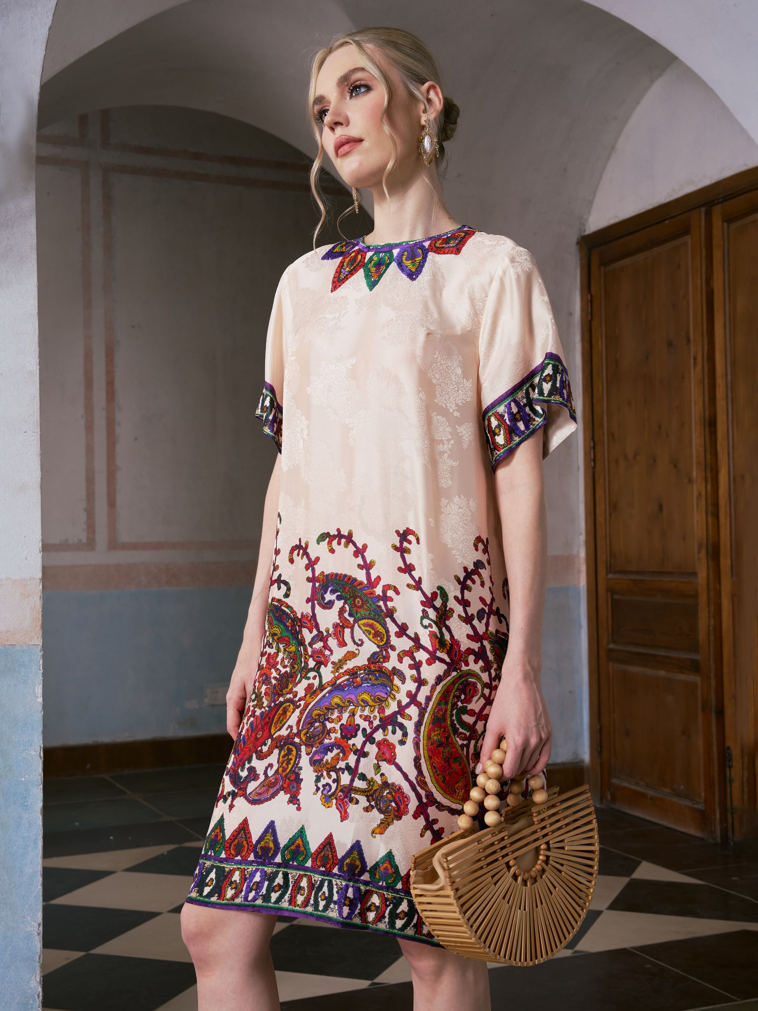 A woman in the Social Queen Paisley Garden Print Jacquard Silk Dress stands indoors, holding a wooden handbag and looking to the side.