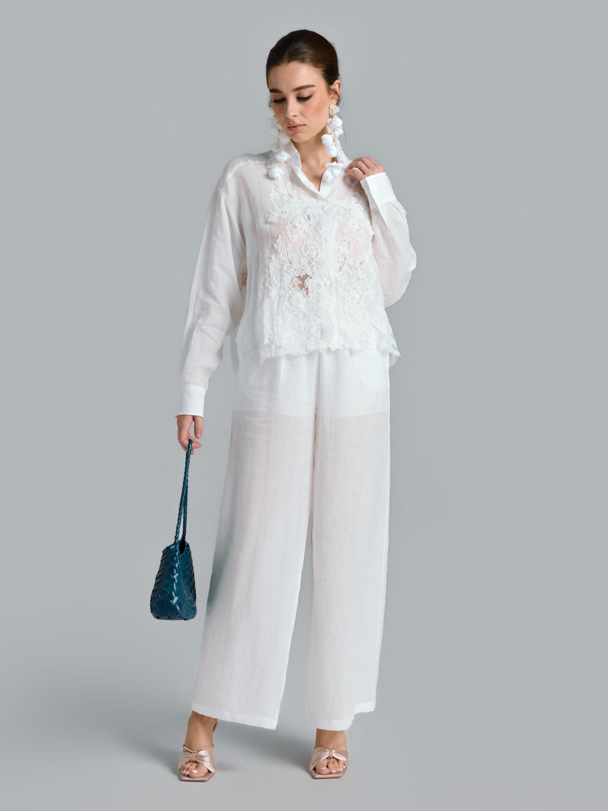 A woman stands against a gray background wearing the Social Queen 3D Lace Appliqué Shirt with sheer white pants, holding a blue purse, and accessorized with large statement earrings and heeled sandals.