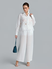 A woman stands against a gray background wearing the Social Queen 3D Lace Appliqué Shirt with sheer white pants, holding a blue purse, and accessorized with large statement earrings and heeled sandals.