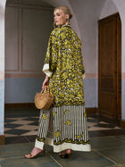 A woman wearing the Social Queen Tropical Fruit Print Linen Dress stands in a hallway, holding a wooden handbag and wearing heeled sandals.