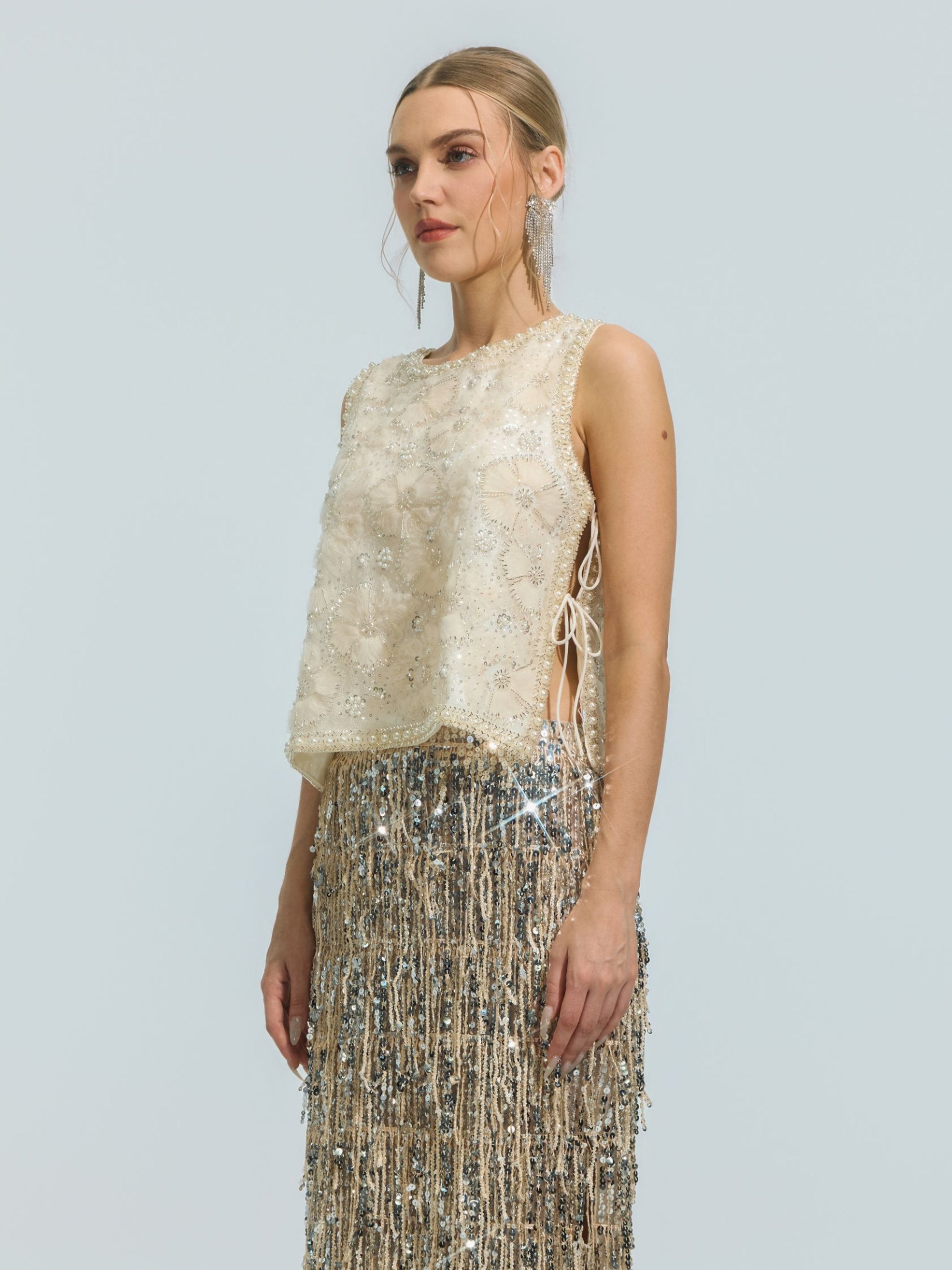 A woman stands against a plain background, looking forward with a neutral expression, wearing the Social Queen Floral Beaded Mesh Sleeveless Top in cream, styled with side ties and paired with a silver fringe skirt adorned with delicate beaded details.