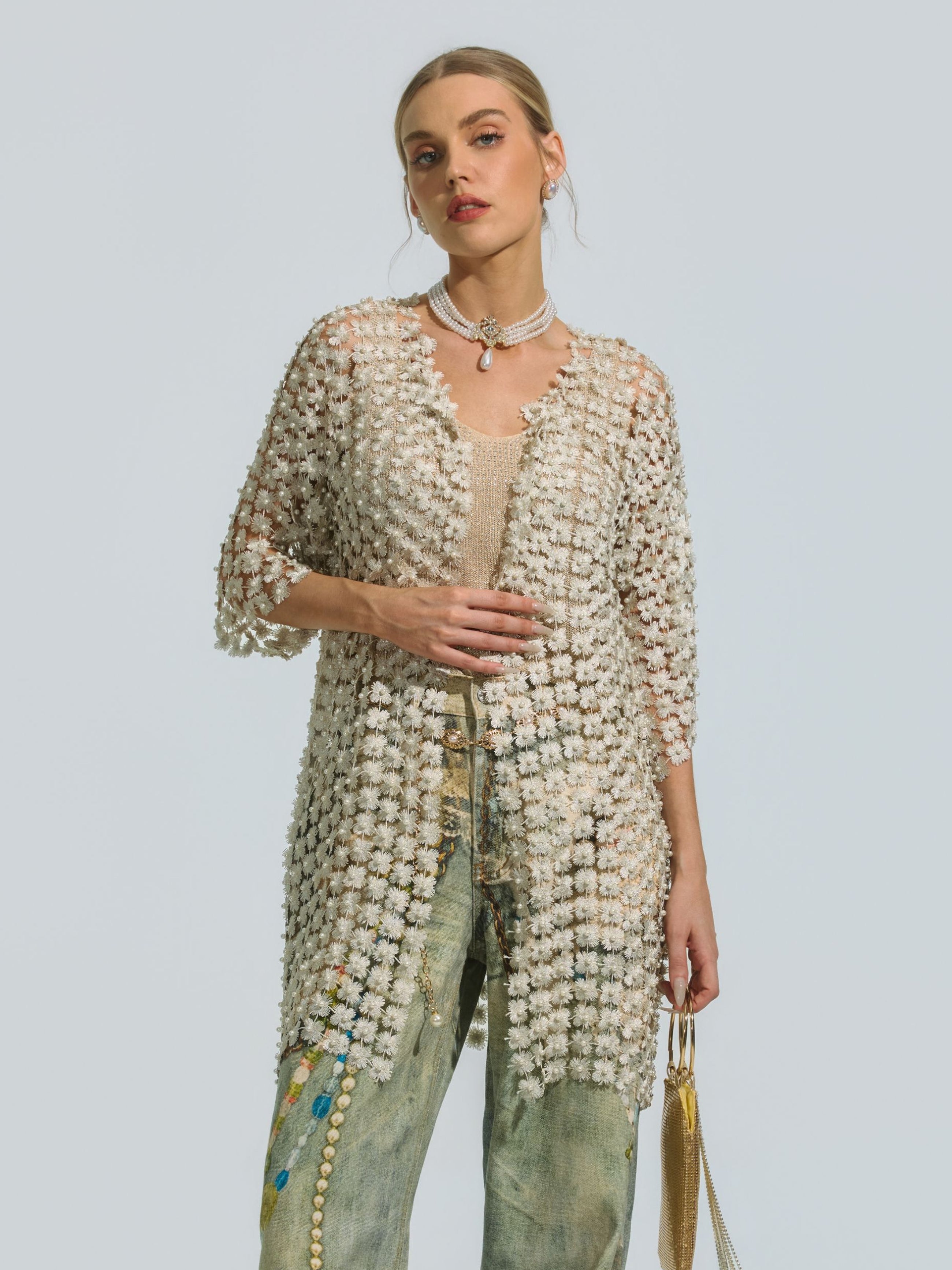 A woman wears the Social Queen Pearl Daisy Cutwork Long Cardigan, featuring delicate pearl accents, over a beige top and jeans. She holds a gold bag against a plain light background, her ensemble elevated by the cardigan’s lightweight polyester design.