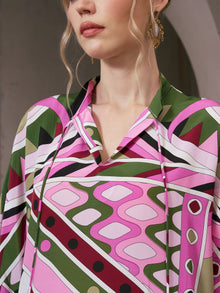A woman stands indoors wearing the Social Queen Geometric Print Mulberry Silk Dress in pink, green, and white hues; only her lower face and upper torso are visible.