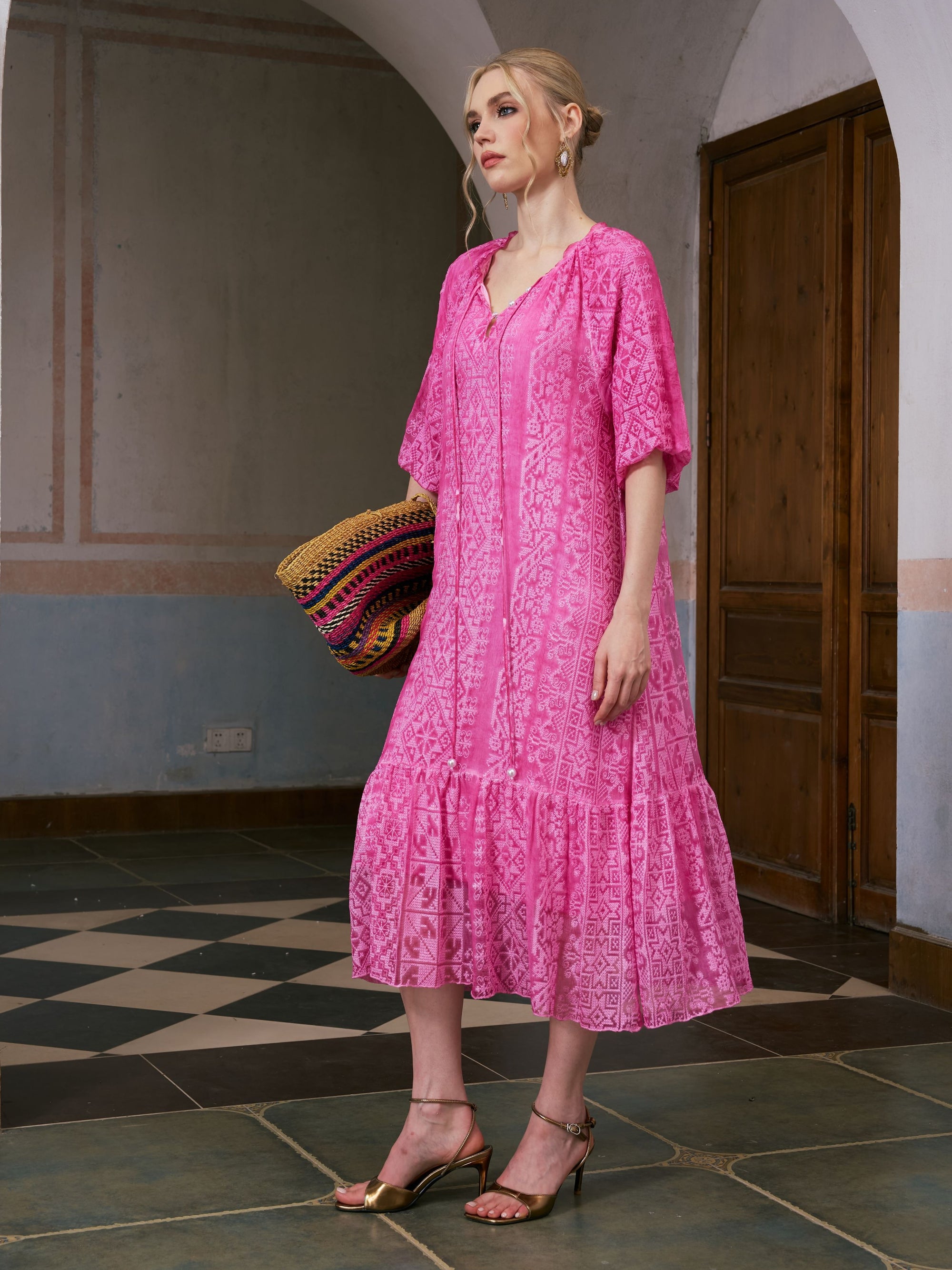 A woman wearing the Social Queen Geometric Jacquard Mulberry Silk Dress and gold sandals stands indoors holding a colorful striped woven bag near a wooden door and tiled floor—perfect for elegant resort wear.