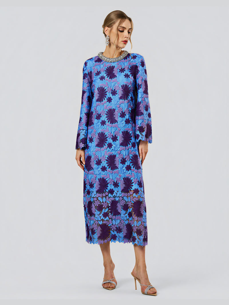 A woman wearing the Social Queen Crystal Trim Lace Shift Dress—a long-sleeved, blue and purple floral midi dress with delicate floral detailing and sheer accents at the hem—stands against a plain light background.
