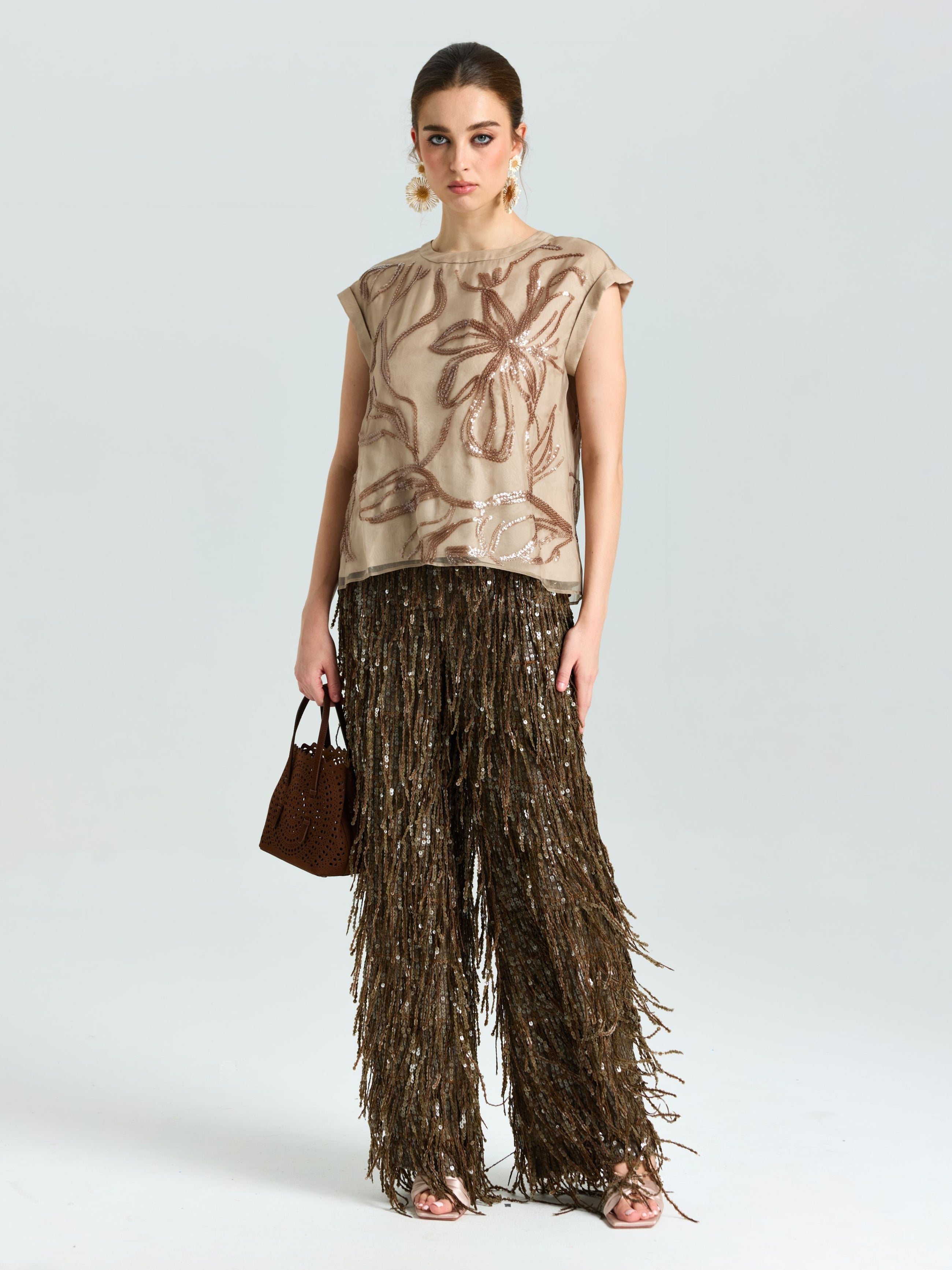 A woman poses against a plain background wearing Social Queen's Sequin Fringe Wide-Leg Pants with a beige abstract-print top, statement earrings, and a small brown handbag—an eye-catching look for evening events.