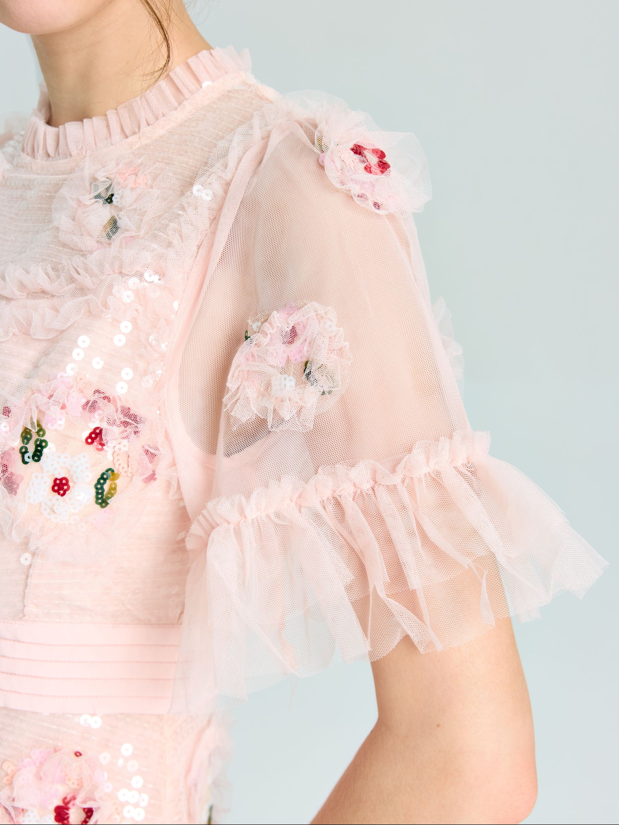 A person wearing the Embroidered Ruffle Tiered Tulle Dress by Social Queen, featuring sheer ruffled sleeves and floral sequin embroidery—an elegant choice for any special occasion.