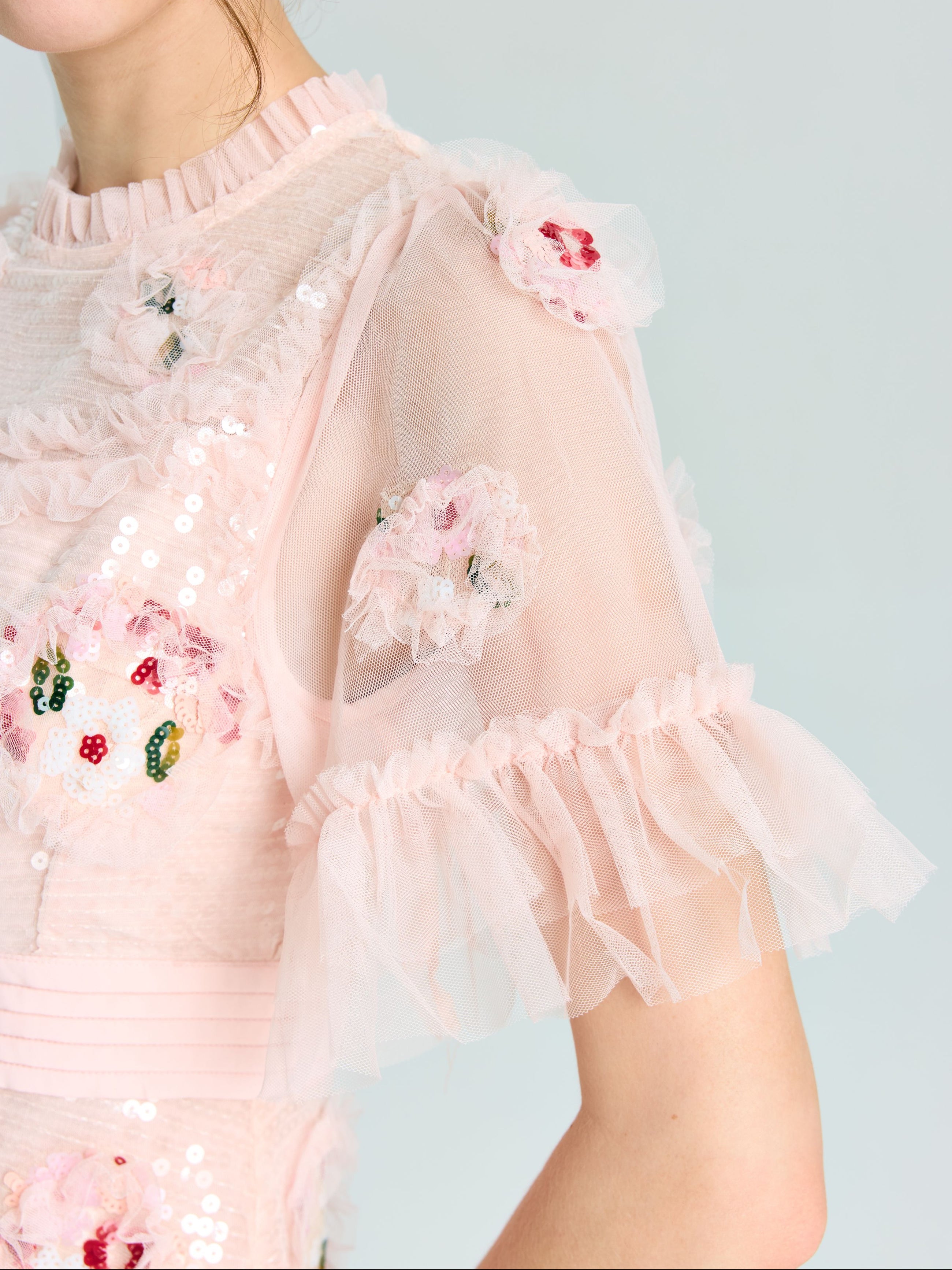 A person wearing the Embroidered Ruffle Tiered Tulle Dress by Social Queen, featuring sheer ruffled sleeves and floral sequin embroidery—an elegant choice for any special occasion.