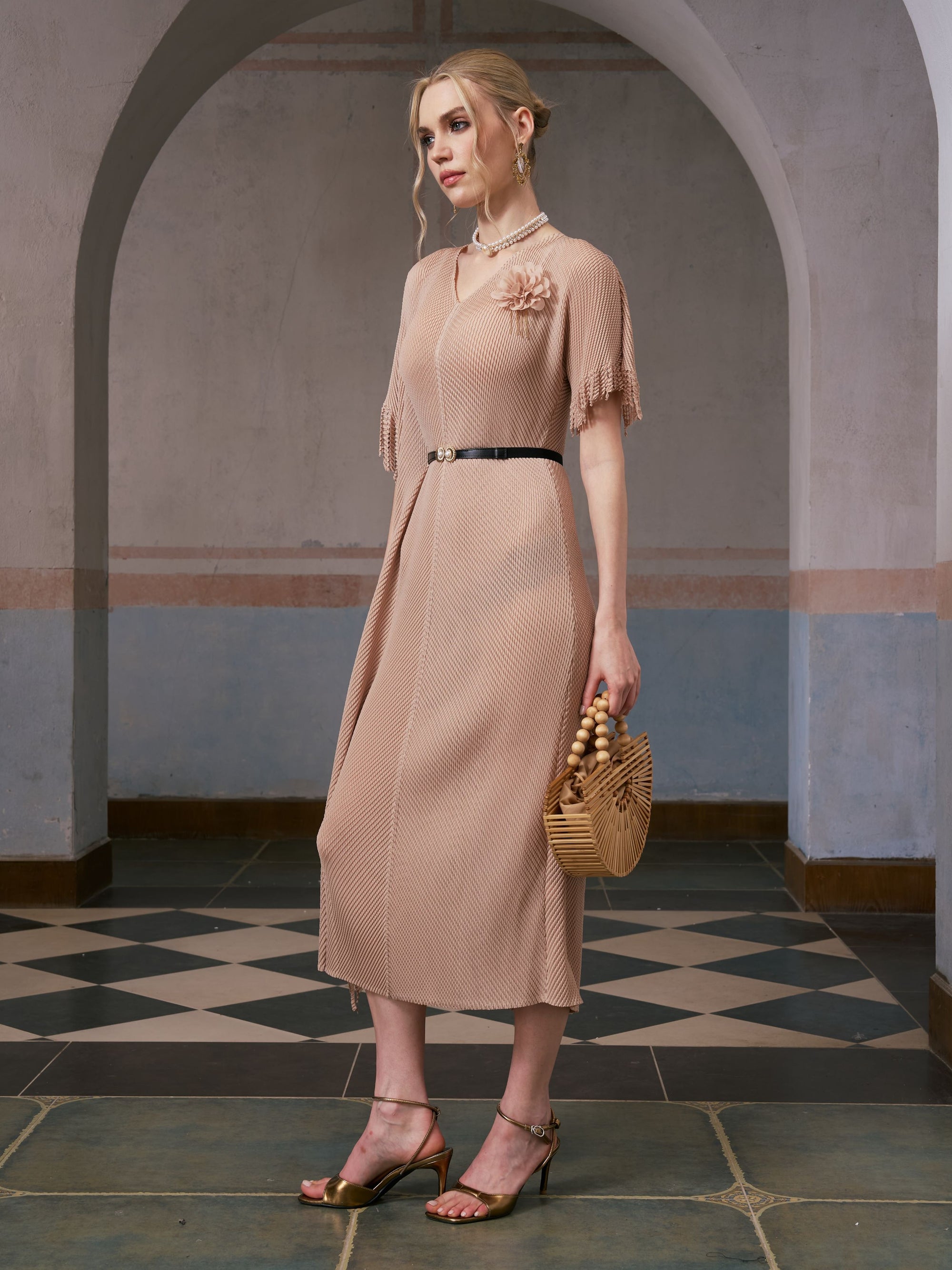 A woman stands in an arched hallway wearing the Social Queen Asymmetric Fringe Midi Dress (belt included), paired with gold heels and a wooden handbag.