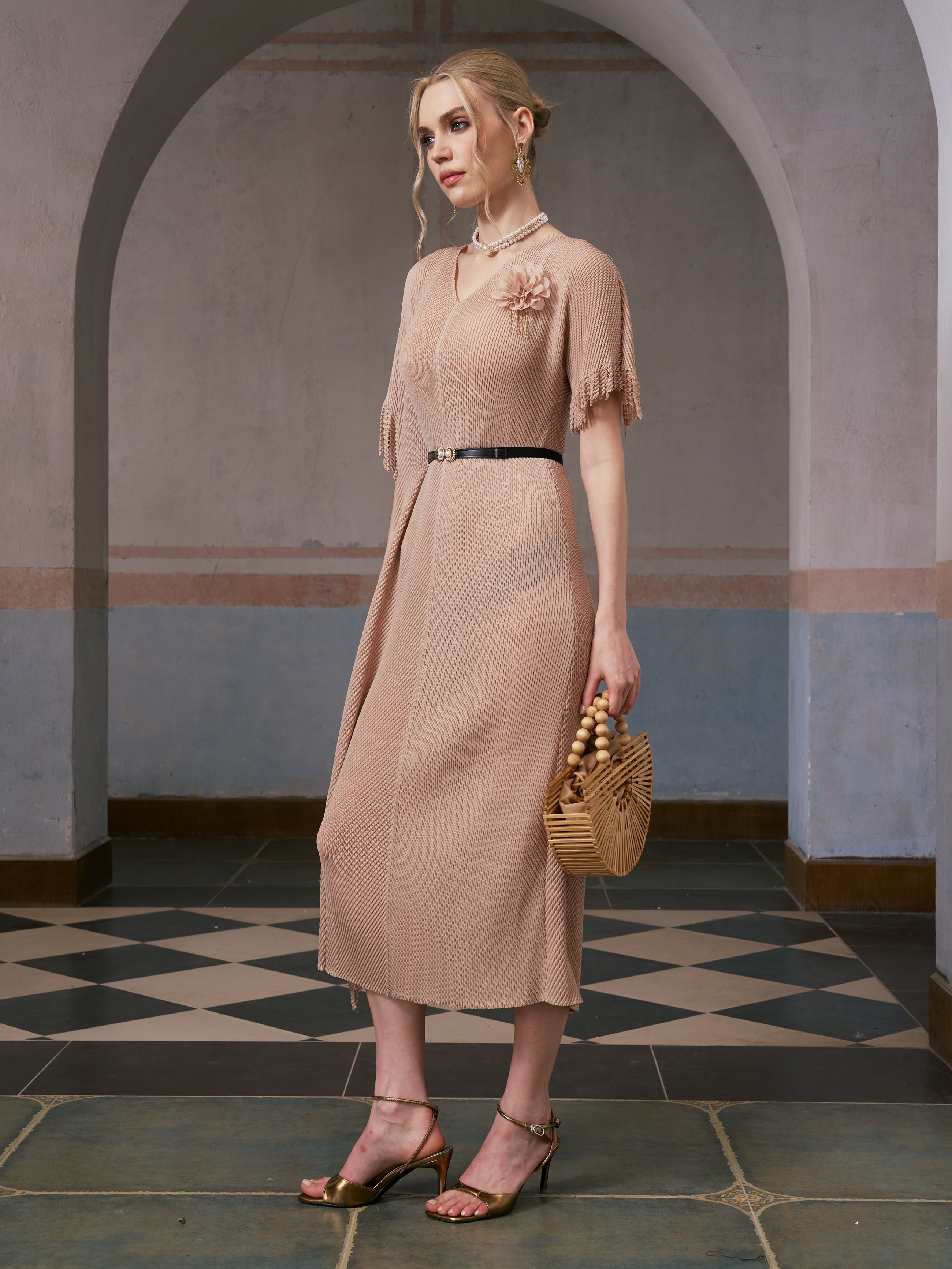 A woman stands in an arched hallway wearing the Social Queen Asymmetric Fringe Midi Dress (belt included), paired with gold heels and a wooden handbag.